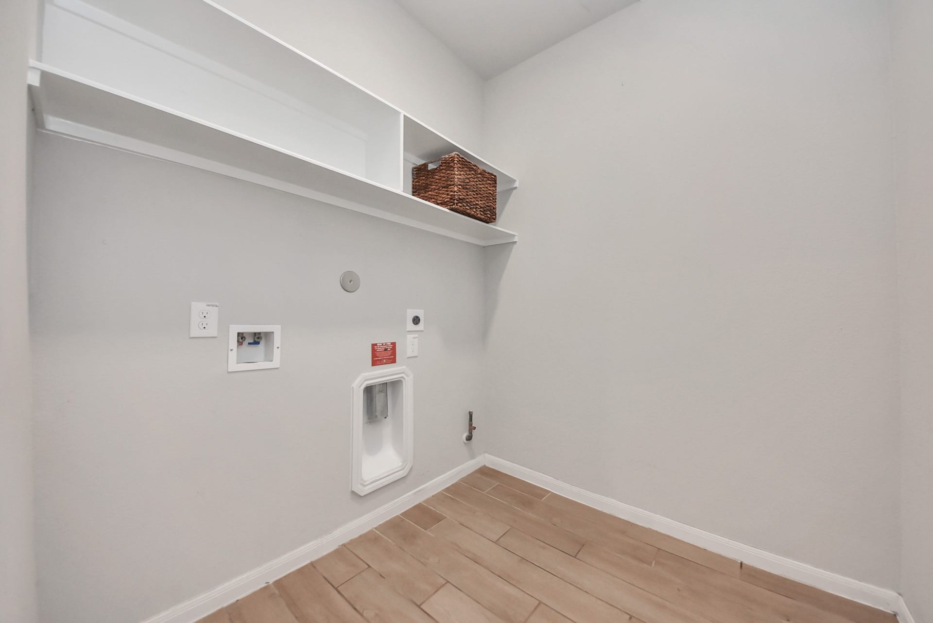 This is a spacious laundry room with light-colored walls and wood-like tile flooring. It features upper shelving for storage and outlets for washer and dryer hookups.