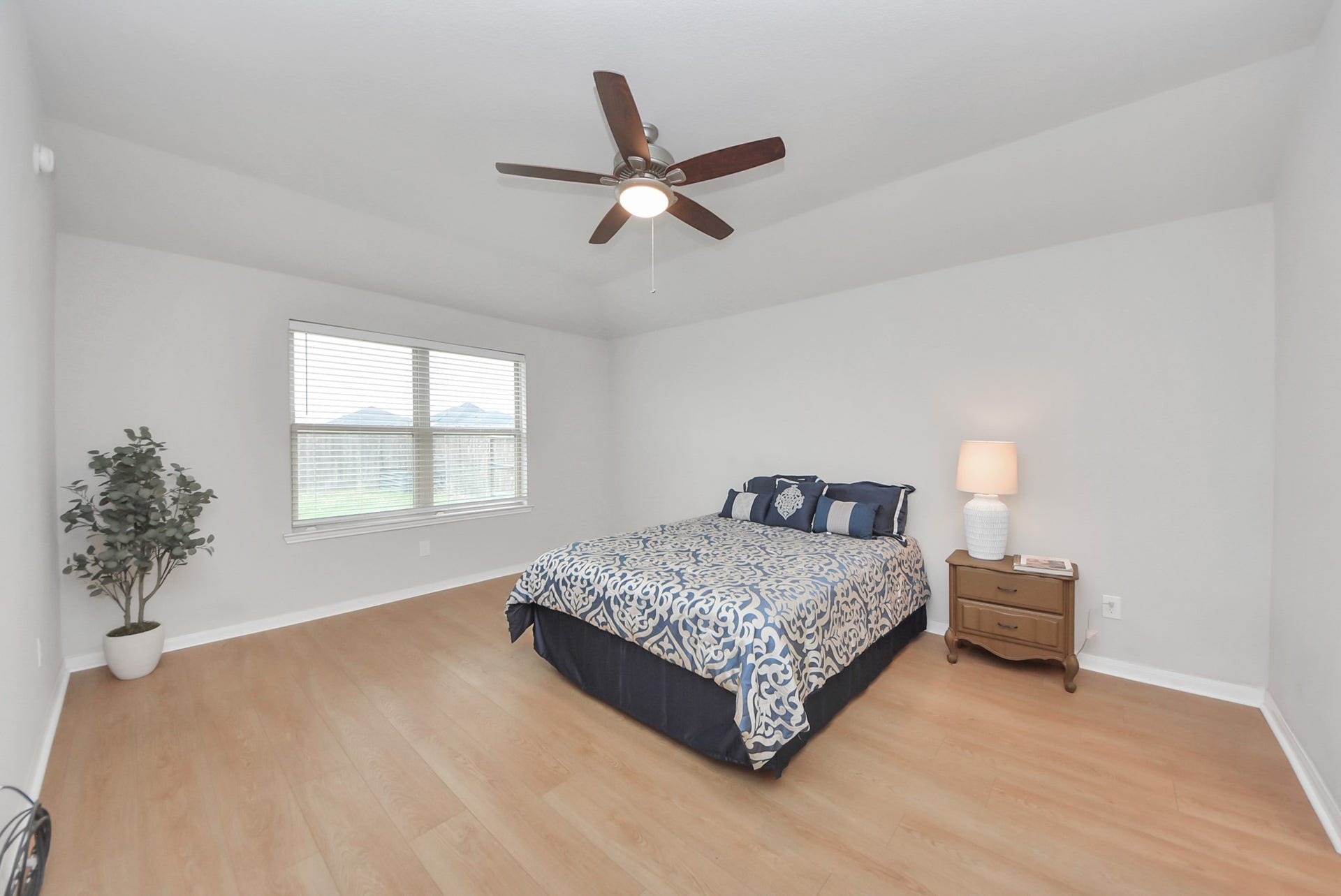 This spacious bedroom features light wood flooring, neutral walls, and ample natural light from a large window. It includes a ceiling fan for comfort, a cozy bed with patterned bedding, and a bedside table with a lamp. A potted plant adds a touch of greenery.