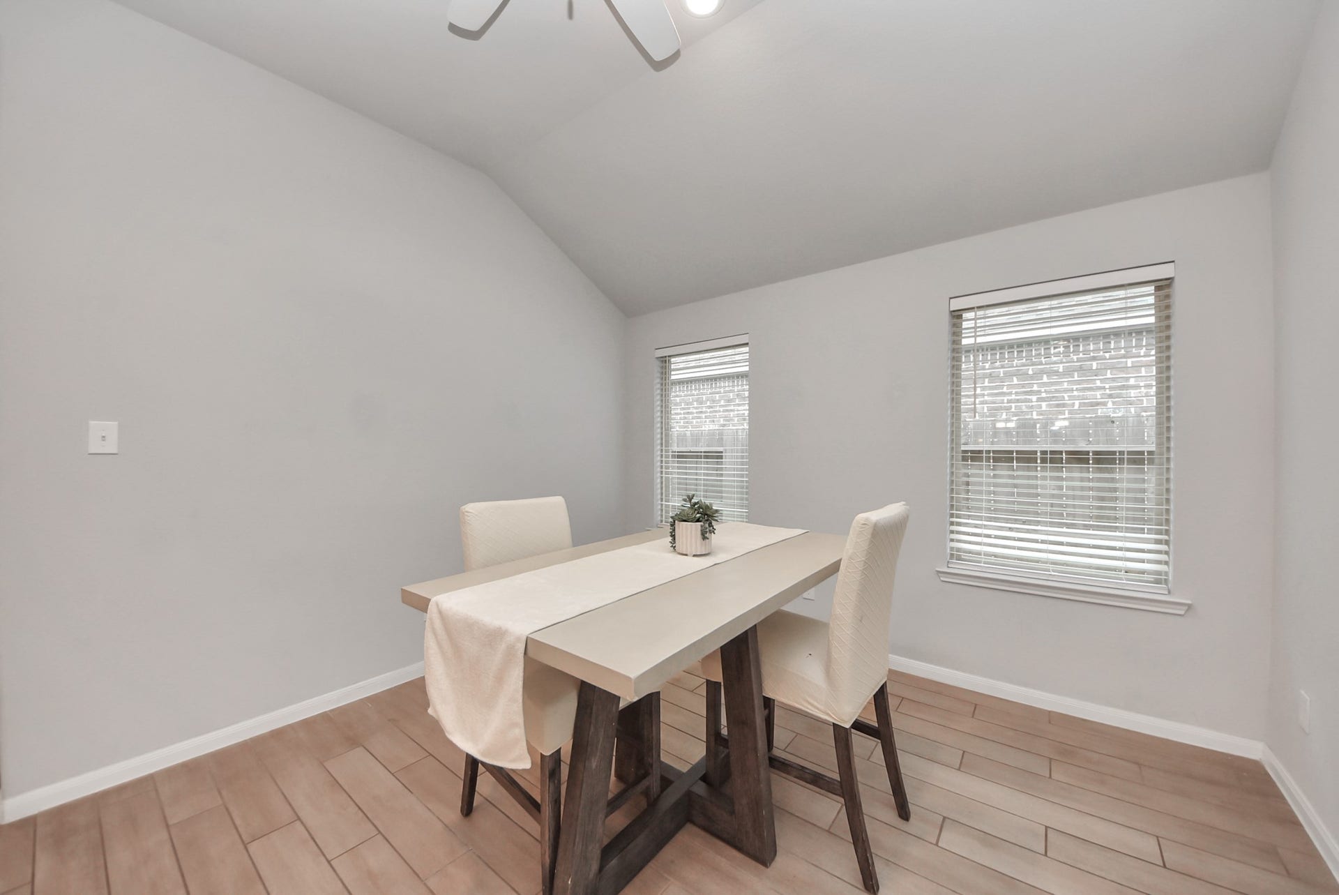 This photo showcases a bright dining area with a vaulted ceiling, featuring a modern table with two chairs. The room has light wood flooring and two windows with blinds, offering a cozy and inviting atmosphere.