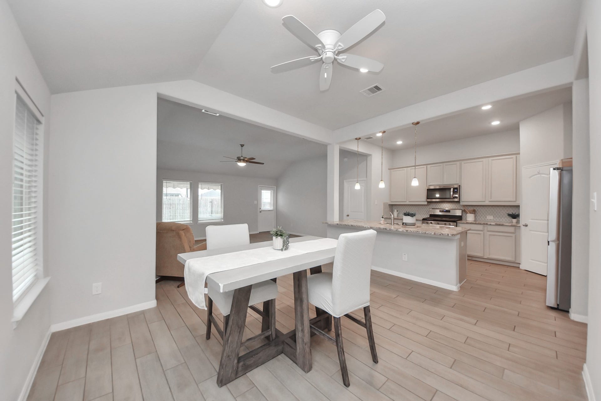 This open-concept kitchen and dining area features modern cabinetry, stainless steel appliances, and a cozy dining space. The wood-look tile flooring and pendant lighting add a stylish touch, while the ceiling fans ensure comfort. The area seamlessly flows into a bright living space with large windows.