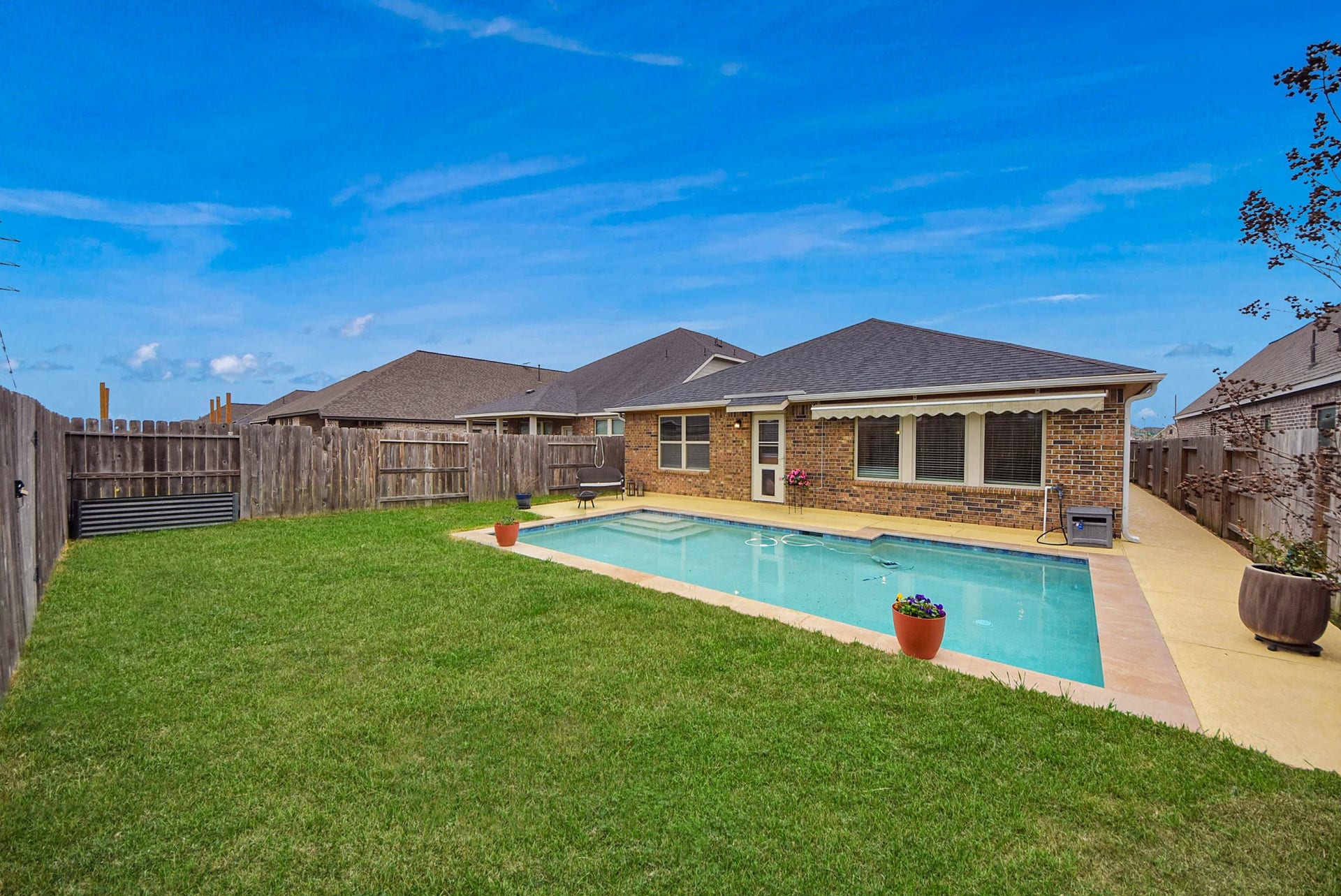 This photo showcases a backyard with a well-maintained lawn and a rectangular swimming pool. The brick house has a patio area, and the yard is enclosed by a wooden fence, providing privacy. Two potted plants add a touch of color to the scene.