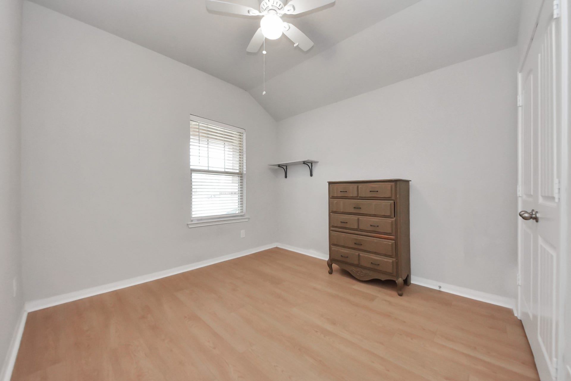 This room features light wood flooring, a neutral color scheme, and a ceiling fan. It includes a window with blinds, a wooden dresser, and a wall shelf, offering a cozy and functional space.
