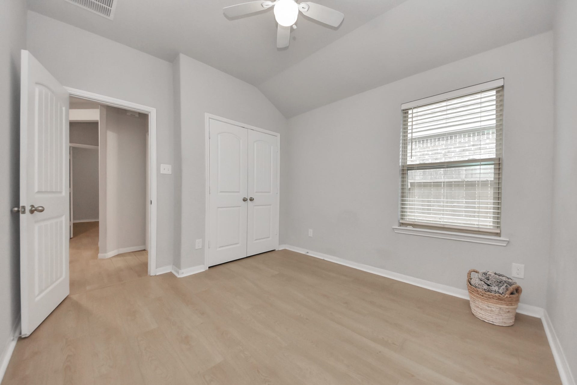 This bright, spacious room features light wood flooring, neutral walls, and a large window with blinds. It includes a ceiling fan and double doors leading to a closet. Ideal for a bedroom or home office.