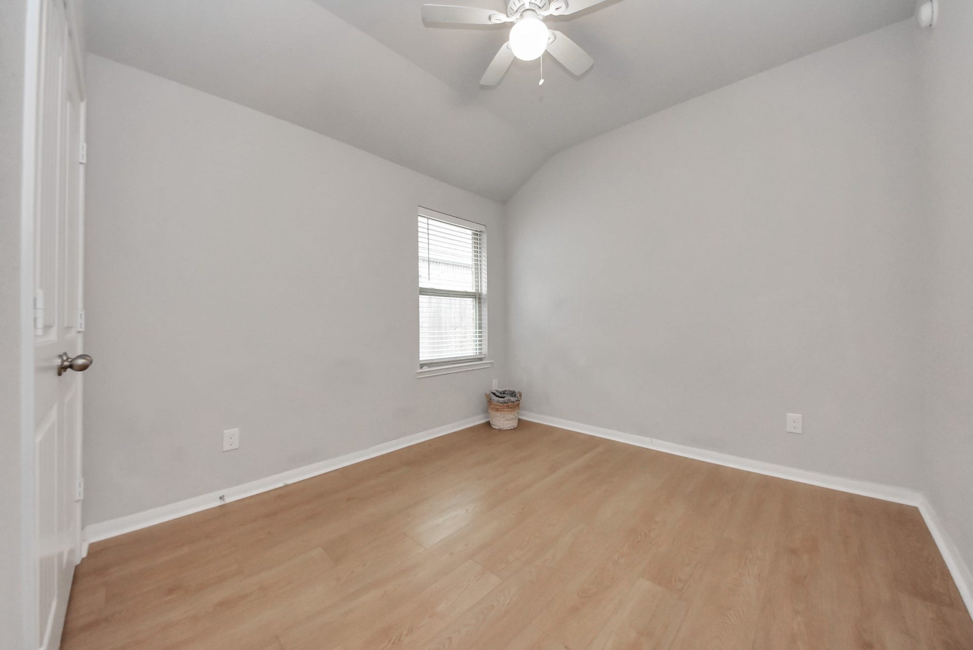 This room features a high ceiling with a ceiling fan, neutral gray walls, and light wood flooring. A window allows natural light, making it a bright and versatile space. Perfect for a bedroom or home office.