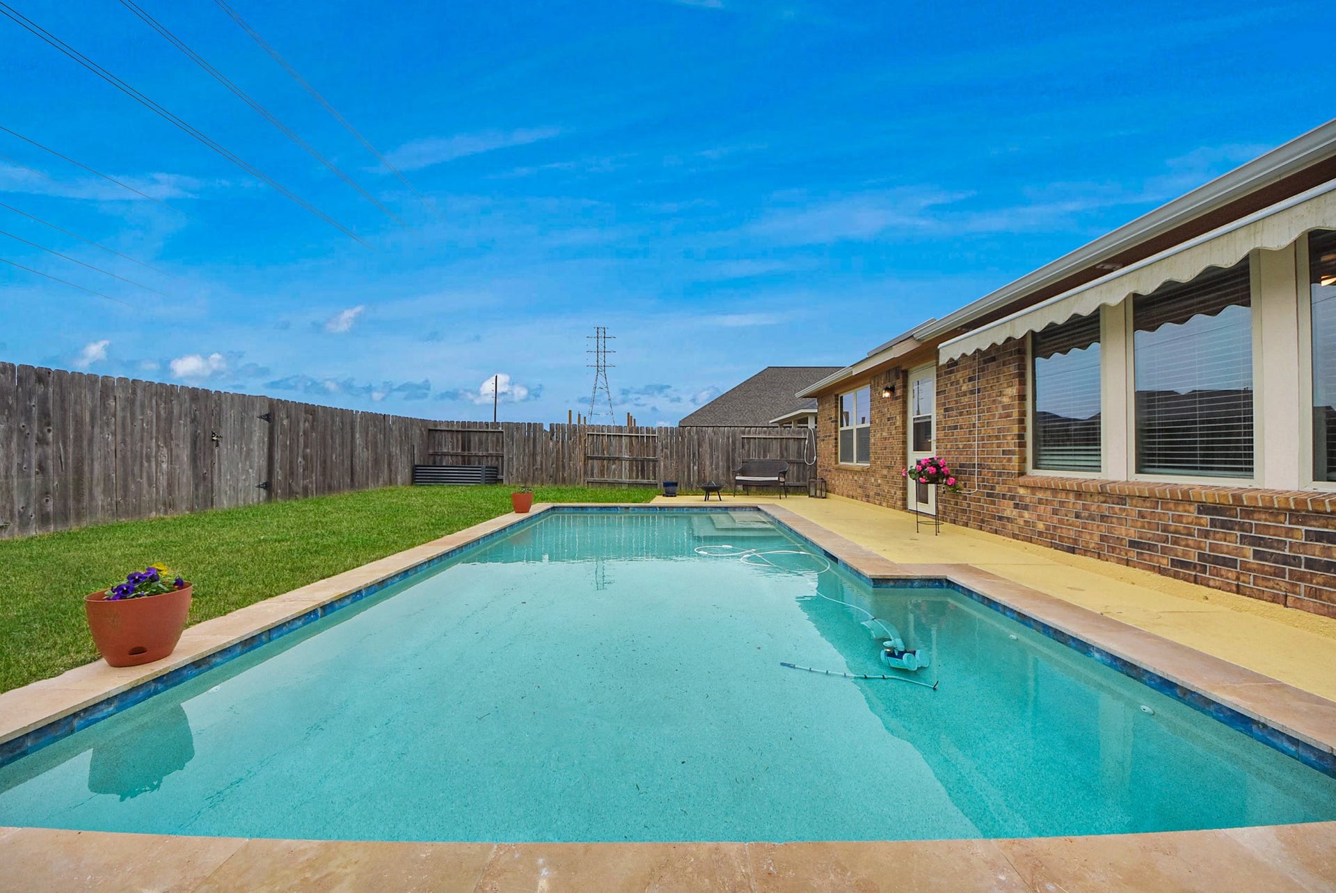 Spacious in-ground pool, bordered by a wooden fence for privacy. The house exterior features brickwork and large windows, and there's a well-maintained lawn with potted plants, ideal for outdoor relaxation.