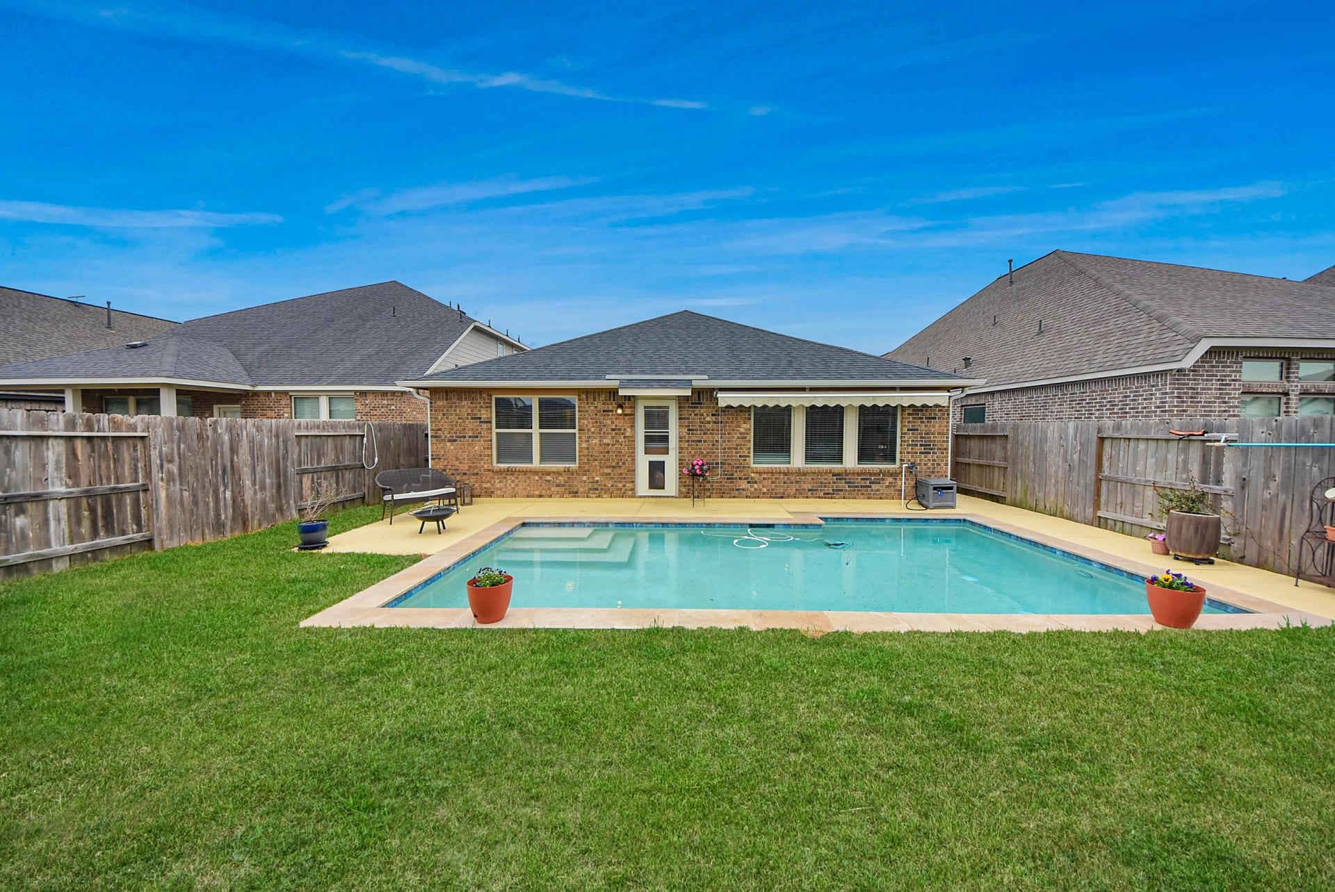 Charming backyard with a spacious in-ground pool, bordered by well-maintained grass and a wooden privacy fence. The brick house features large windows and a cozy patio area, perfect for relaxing or entertaining.