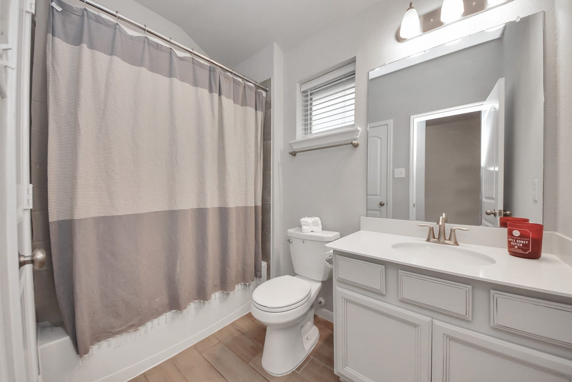 bathroom features a clean, modern design with light wood flooring. It includes a shower with a neutral-toned curtain, a white vanity with ample storage, a large mirror, and a window for natural light.
