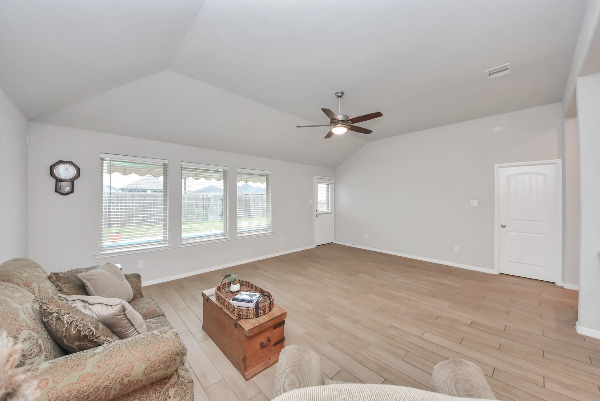 Bright and spacious living room with vaulted ceiling, light wood flooring, and large windows offering ample natural light. Features a ceiling fan and neutral walls, creating a welcoming ambiance.