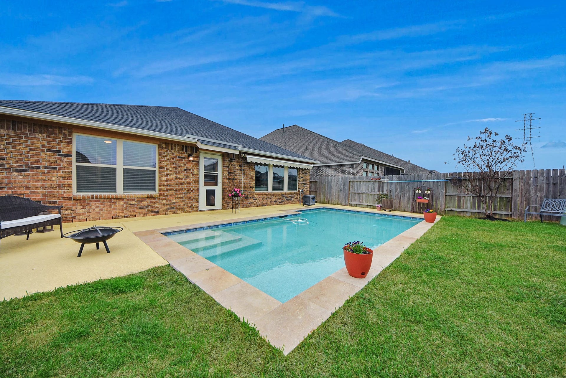 Our backyard features a charming brick house with a well-maintained in-ground pool, surrounded by a concrete patio and lush grass. The area is enhanced with potted plants, a cozy seating bench, and a wooden fence for privacy. Perfect for relaxation and outdoor entertainment.