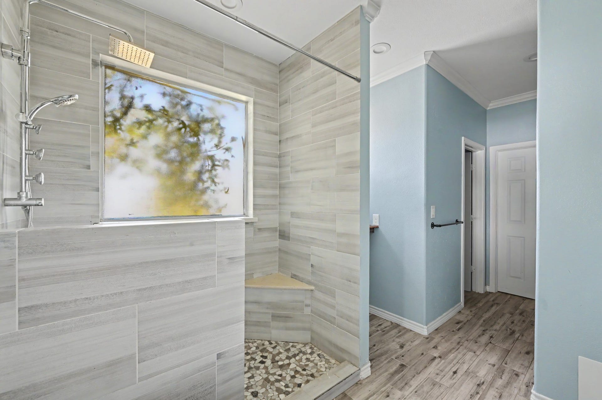 Spacious walk-in shower with floor to ceiling tile, built-in bench, pebble-style shower floor, and natural light from the window.