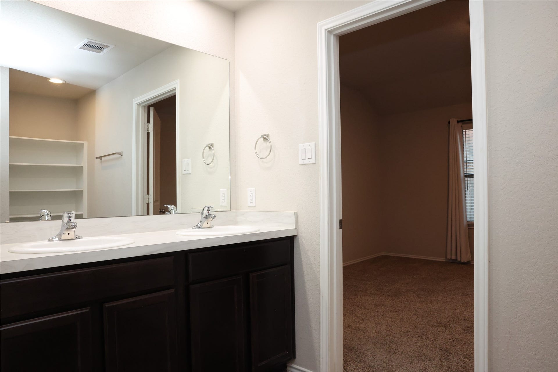 Elegant bathroom with dual sinks and rich dark cabinetry, leading into a cozy carpeted bedroom with natural light.