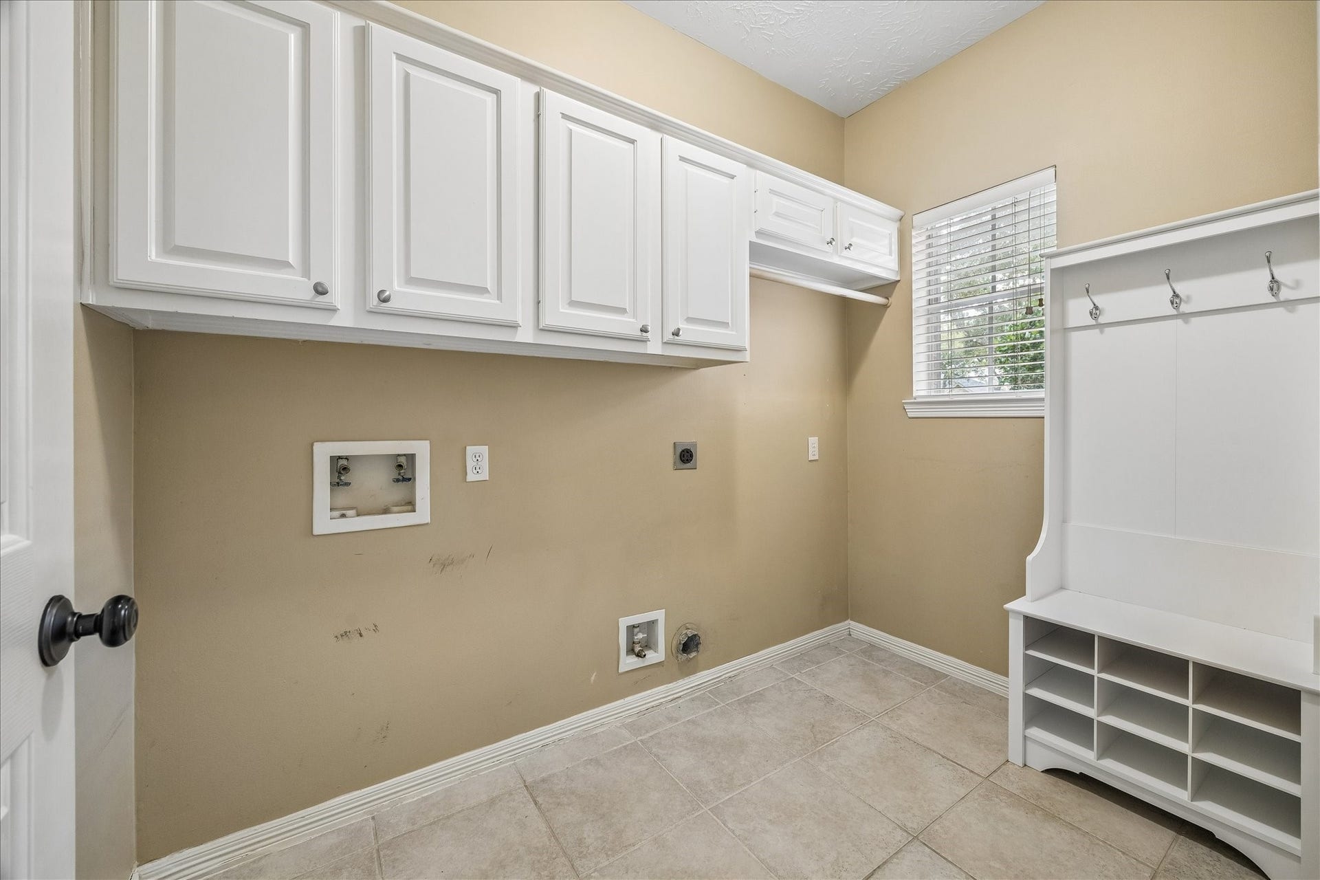 5310 Summerland Ridge Court, Houston, TX 77041 This is a bright laundry room with beige walls and white cabinetry. It features hookups for a washer and dryer, a window for natural light, and a white storage bench with hooks and cubbies.