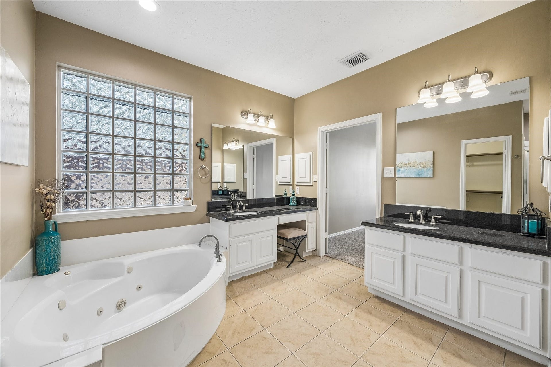 5310 Summerland Ridge Court, Houston, TX 77041 This spacious bathroom features a luxurious corner jacuzzi tub, dual sinks with granite countertops, and a vanity area. It has ample natural light from a large glass block window, elegant tile flooring, and modern light fixtures, creating a serene and functional space.