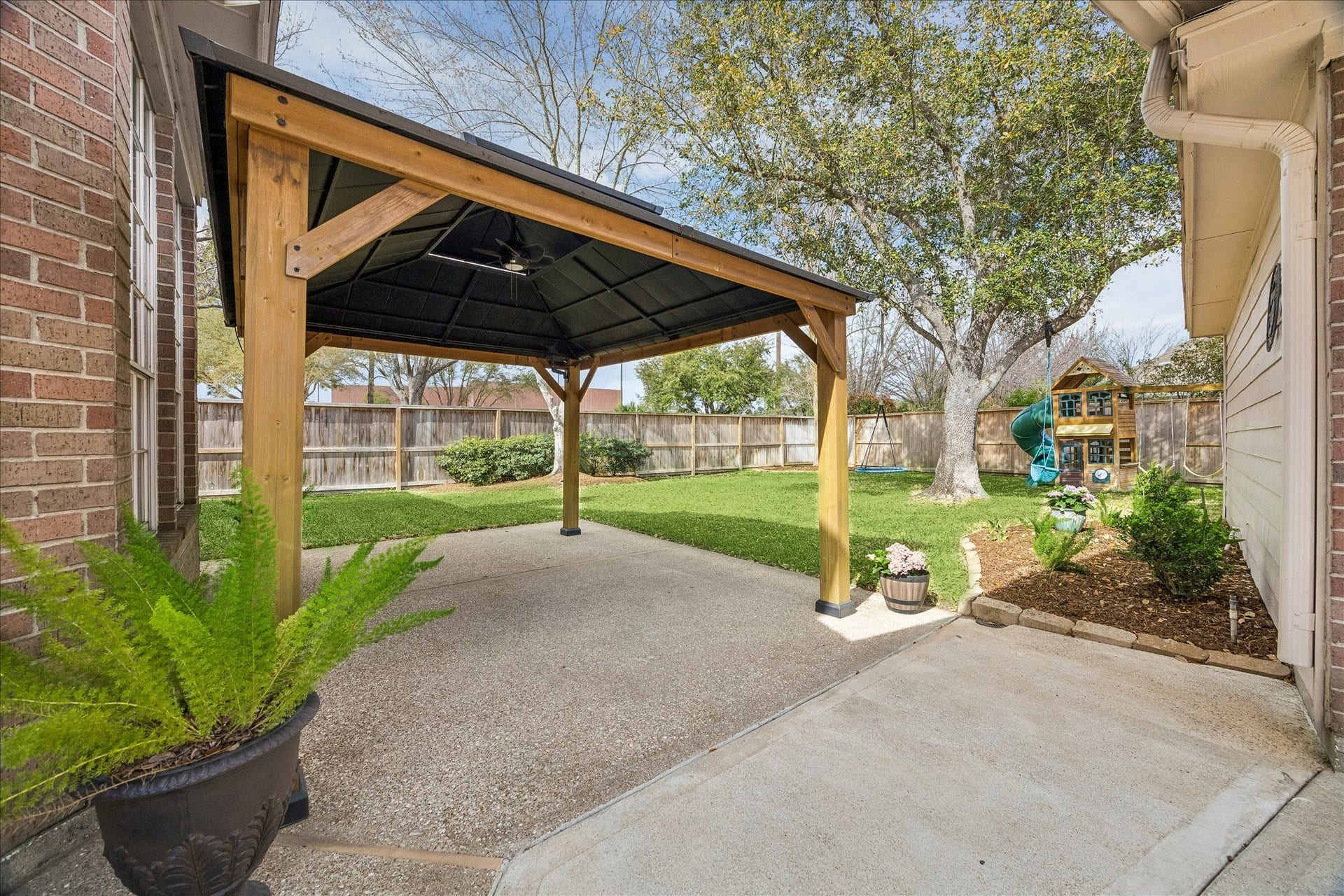 5310 Summerland Ridge Court, Houston, TX 77041 This backyard features a spacious covered patio with a wooden pergola, perfect for outdoor gatherings. The lawn is surrounded by a wooden fence for privacy, and includes a charming children's playset under a large tree. An inviting space for relaxation and family fun.