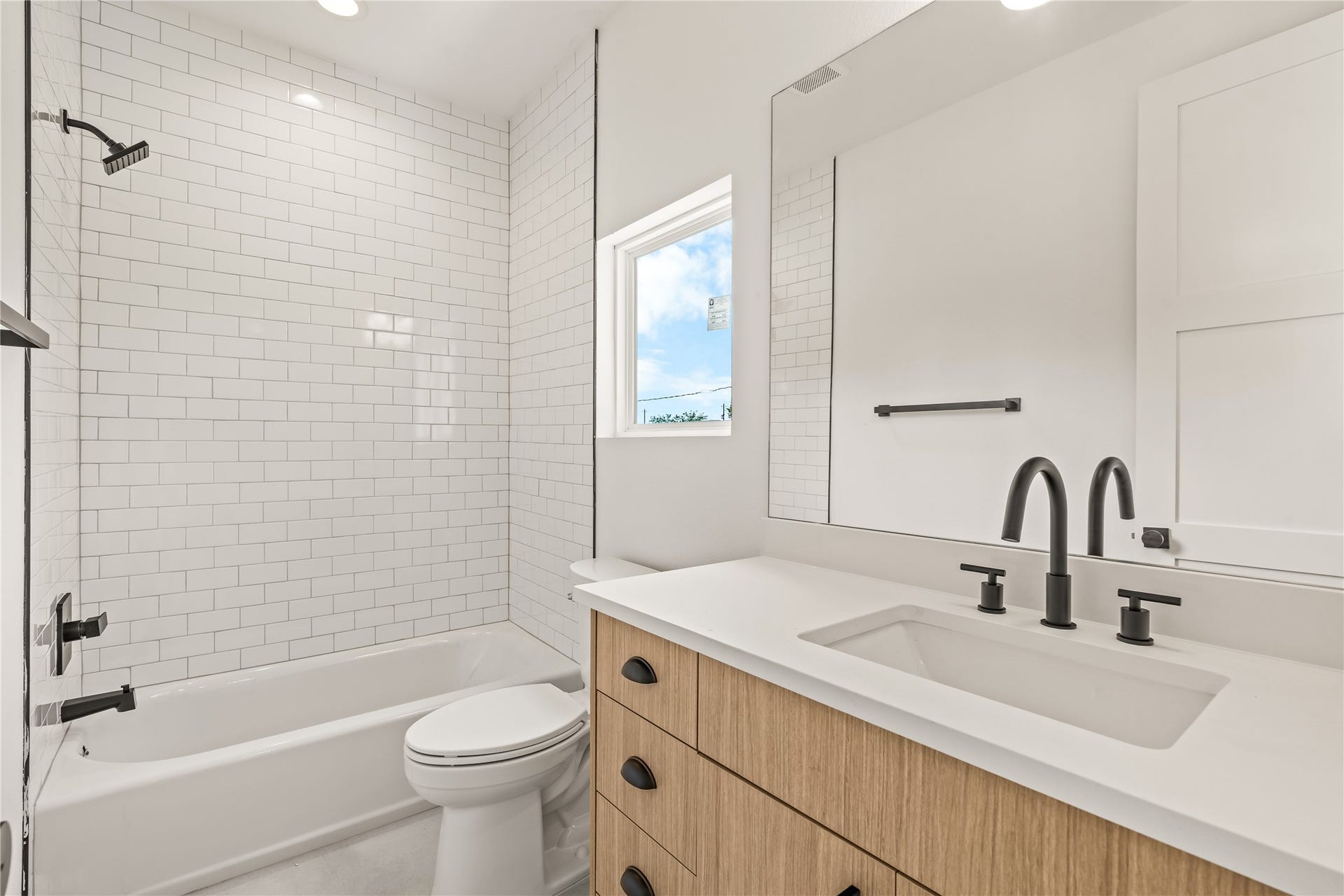 7432 & 7444 Tremper Street, Houston, TX 77020 Modern guest bathroom featuring a light wood vanity with a white countertop, matte black hardware, a large frameless mirror, and a classic white subway-tiled tub/shower combination.