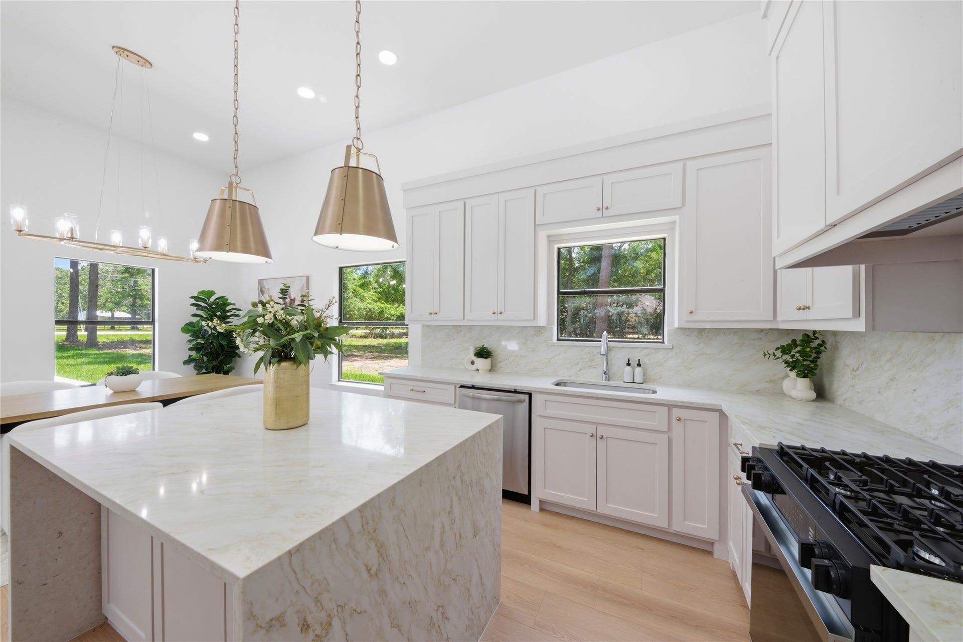 40203 Cimarron Way, Magnolia, TX 77354 Light-filled kitchen showcasing dual brass pendants over the waterfall quartz island, statement linear chandelier above breakfast nook, walls of windows overlooking wooded acreage, and custom cabinetry with brass hardware.