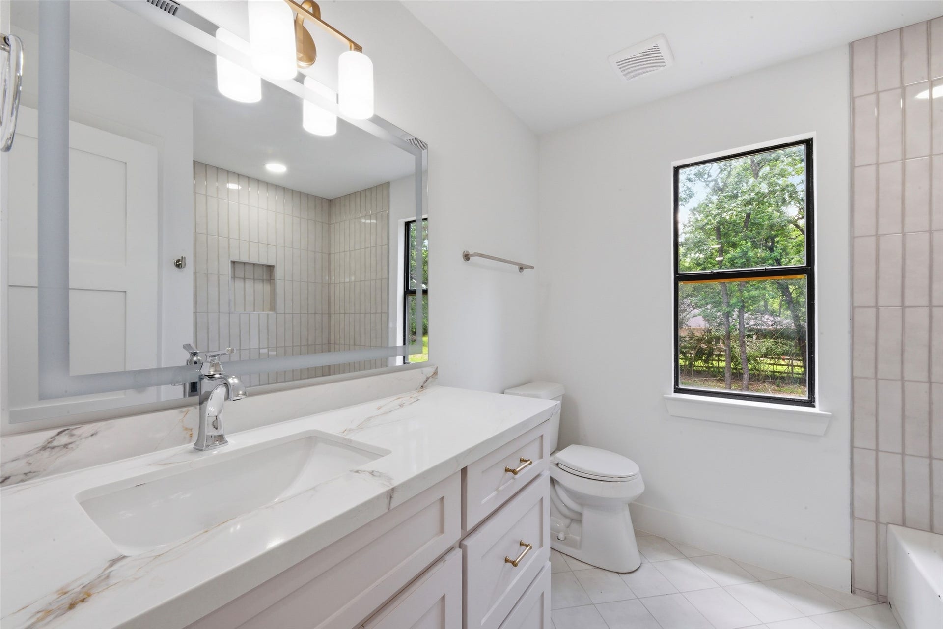 40203 Cimarron Way, Magnolia, TX 77354 Designer secondary bath featuring quartz counter with marble veining, brass sconces, vertical tile tub/shower surround with niche, and natural light from oversized window. Brass hardware throughout.
