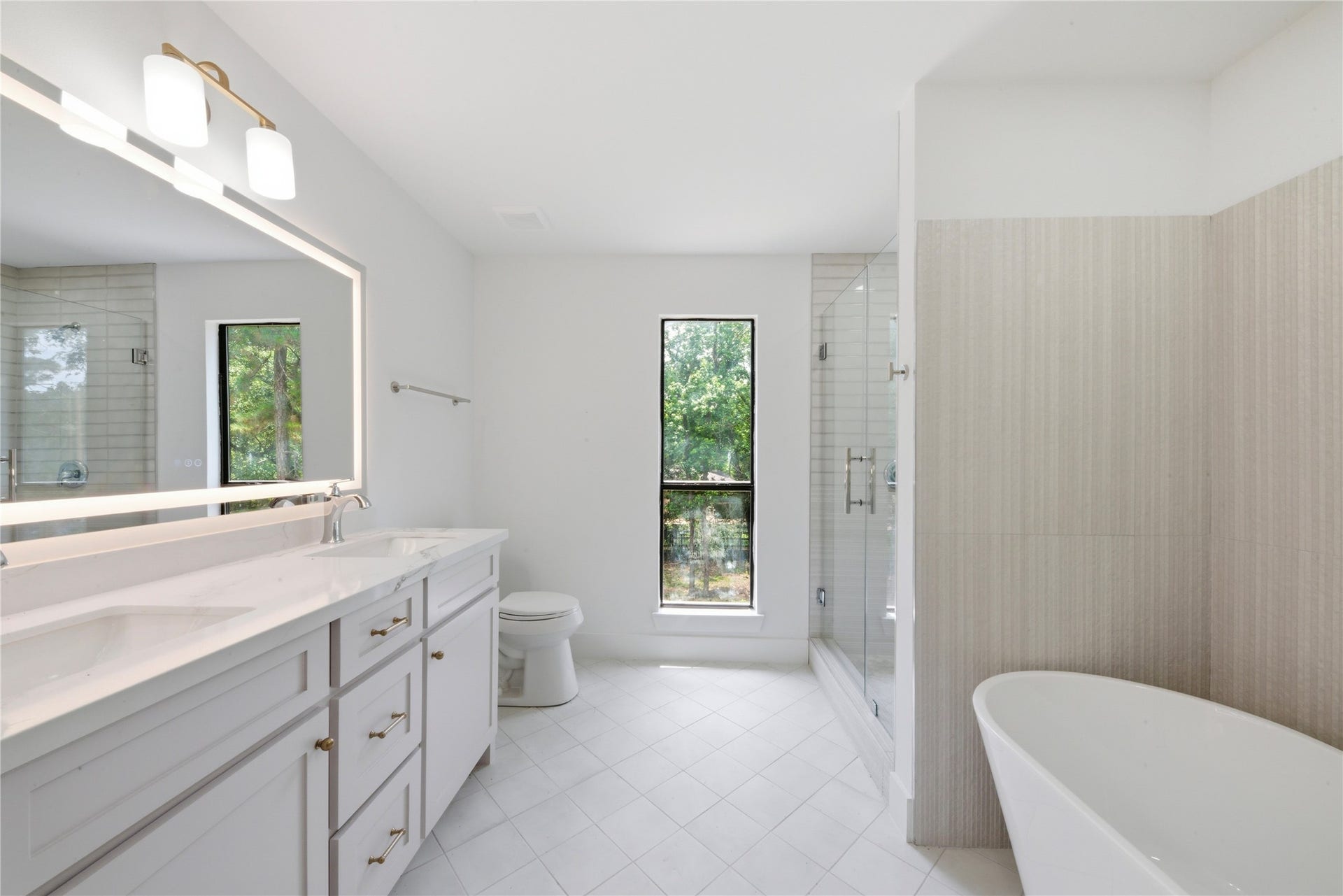 40203 Cimarron Way, Magnolia, TX 77354 Luxurious fourth-floor ensuite bath serving the second primary suite. Features dual quartz vanity, LED-backlit mirror, brass fixtures, freestanding soaking tub, frameless glass shower with vertical tile, and serene wooded views.