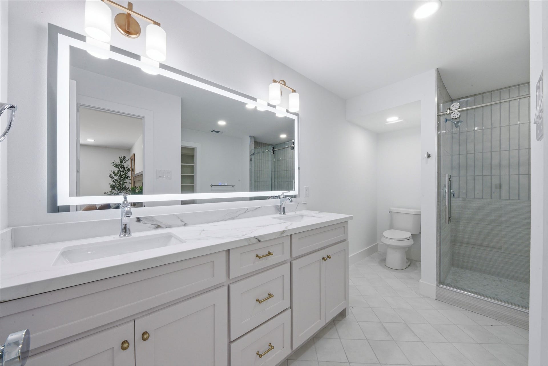 40203 Cimarron Way, Magnolia, TX 77354 Spa-style primary bath featuring dual vanity with quartz counters, LED-backlit designer mirror, brass sconces, large frameless walk-in shower with vertical tile, and separate water closet. Marble-look tile flooring.