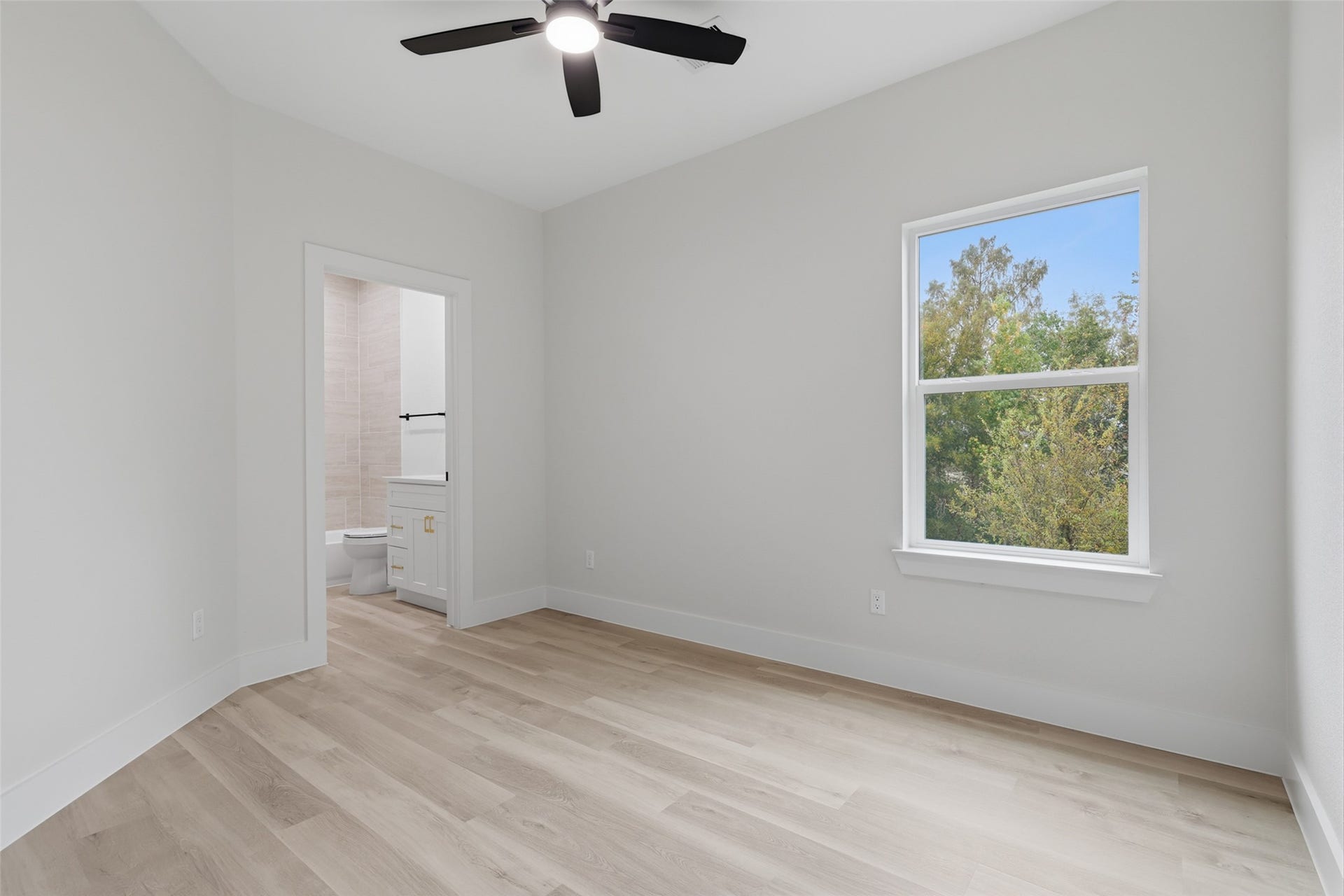 Bright and airy bedroom featuring sleek LVP floors, a ceiling fan, and a large window with a view of lush greenery, leading to a modern en-suite bathroom.