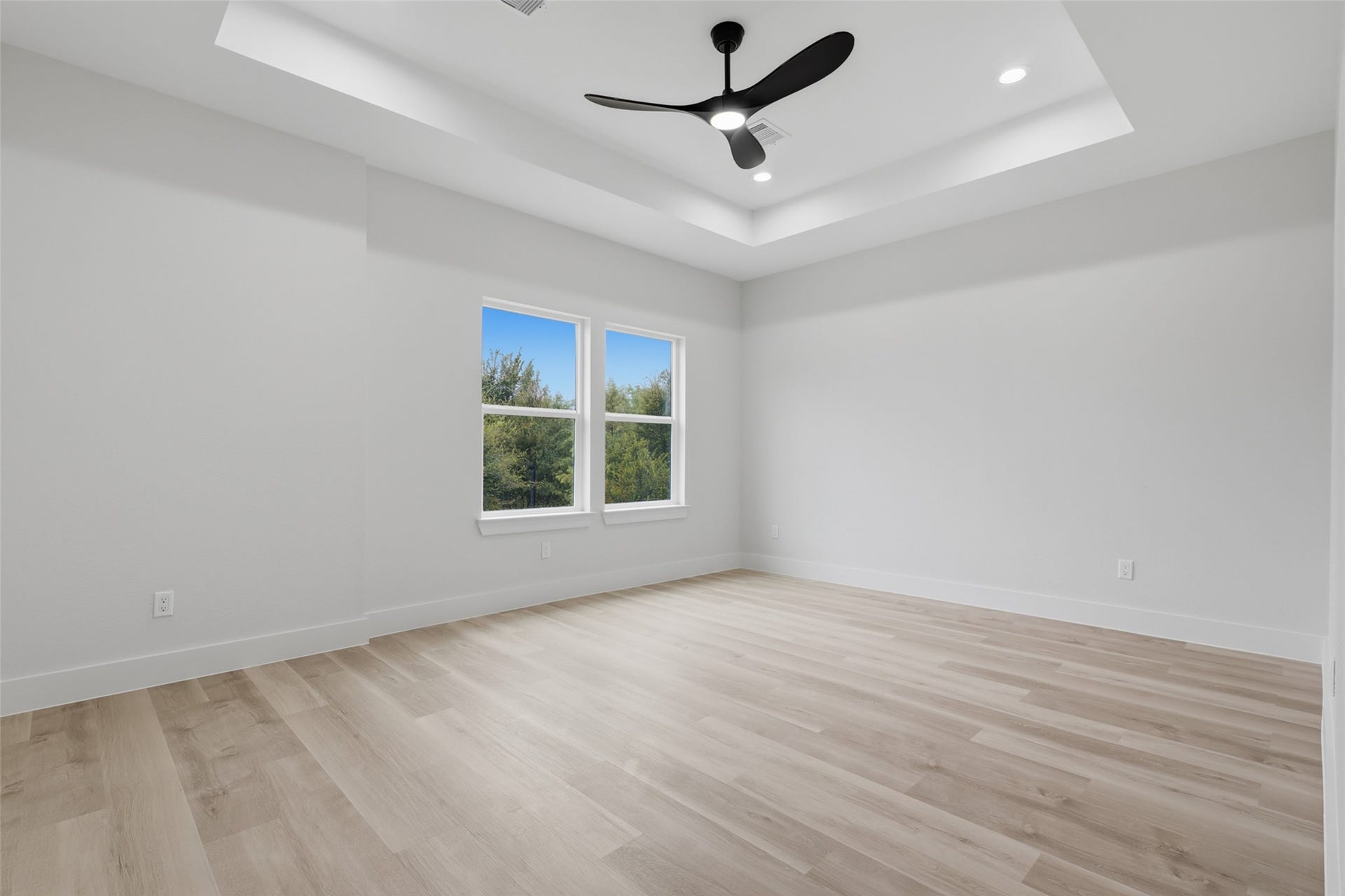 Spacious primary bedroom featuring a tray ceiling with modern fan, abundant natural light from large windows, and sleek LVP flooring.