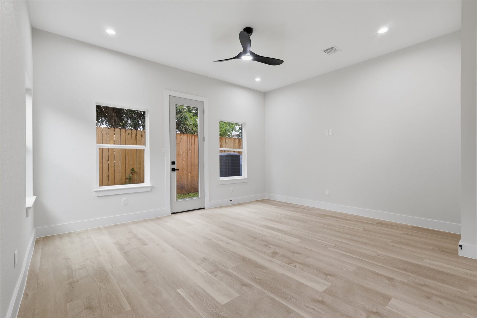 Spacious and bright room featuring sleek LVP floors, modern recessed lighting, and a ceiling fan, with large windows and a glass door leading to a fenced backyard.