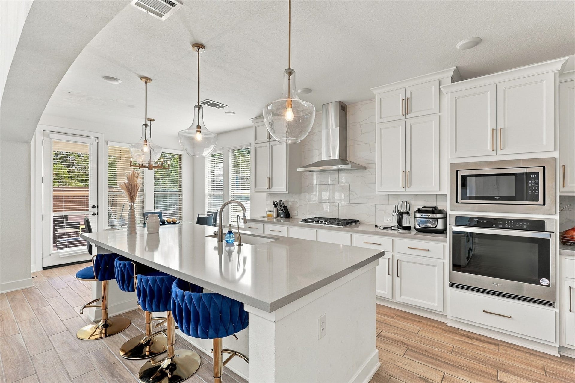 2922 Opal Ivory Way, Richmond, TX 77406 Modern kitchen with sleek white cabinetry, stainless steel appliances, and a spacious island featuring chic blue velvet stools. Ample natural light pours through large windows, highlighting the elegant pendant lighting and contemporary design.