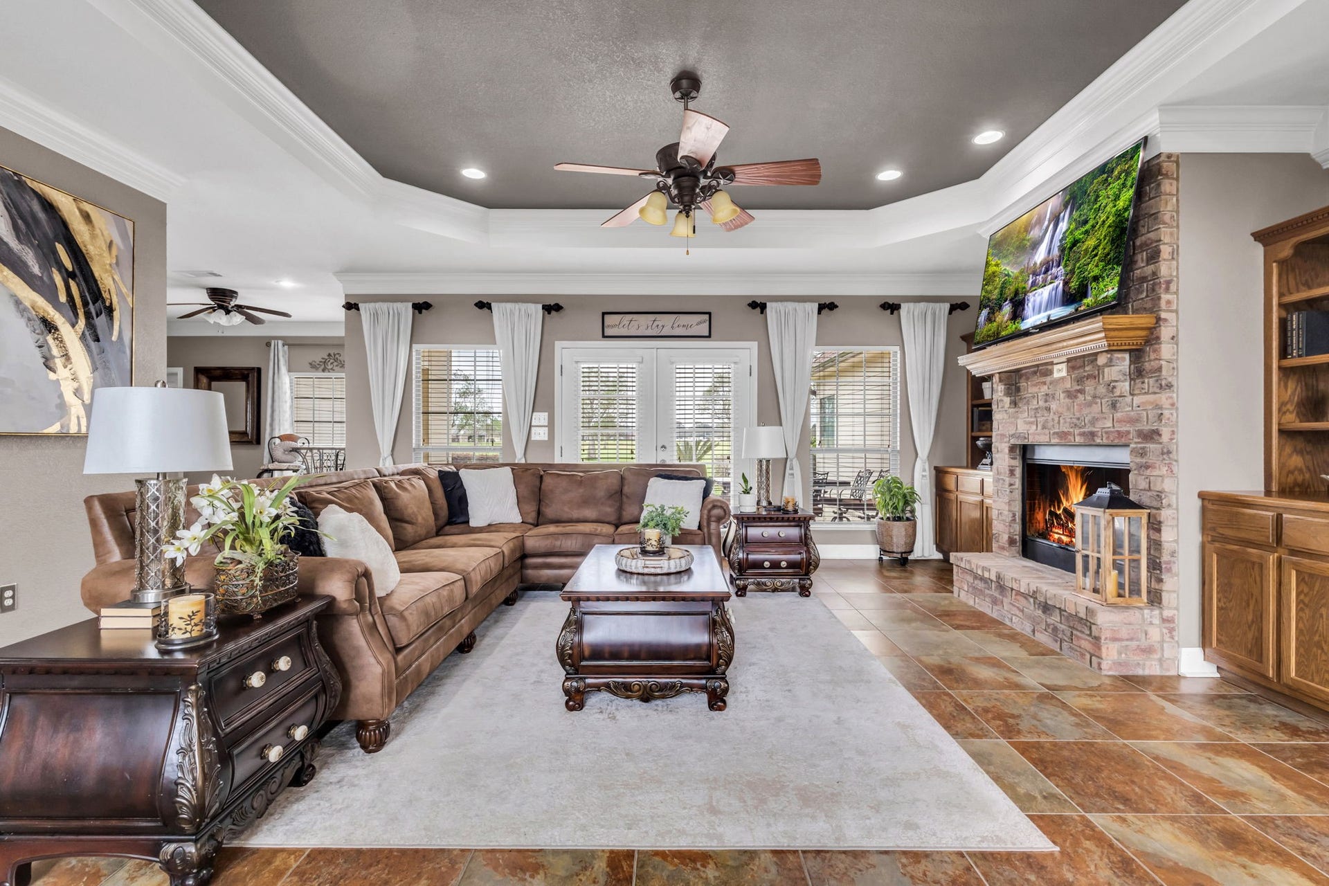11010 Madison Court, Beaumont, TX 77705 Another view of the spacious living room showcasing the floor-to-ceiling brick fireplace, custom built-in cabinetry, and tray ceiling with crown molding. French doors and surrounding windows offer plenty of natural light and views of the backyard.