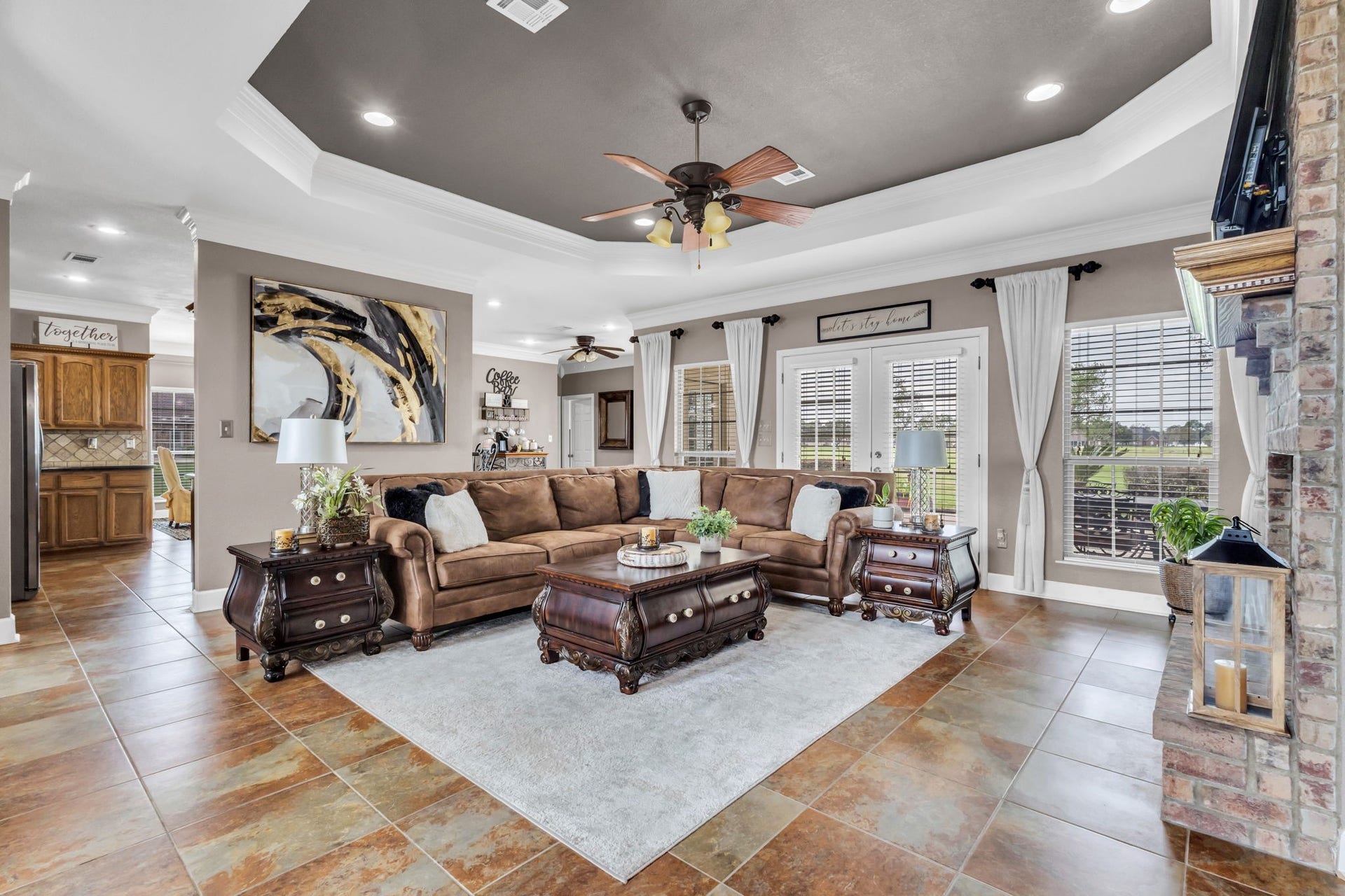 11010 Madison Court, Beaumont, TX 77705 Spacious living room featuring a tray ceiling with crown molding, recessed lighting, tile flooring, and a brick fireplace. Large windows and French doors provide natural light, with an open layout connecting to the kitchen and breakfast area.