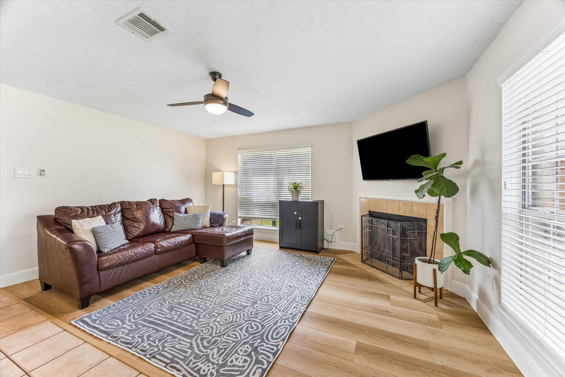 1043 Danforth Drive, Houston, TX 77062 Bright and inviting living room featuring a cozy leather sectional, modern ceiling fan, and a stylish corner fireplace. Abundant natural light flows through large windows, highlighting the elegant flooring and contemporary decor.