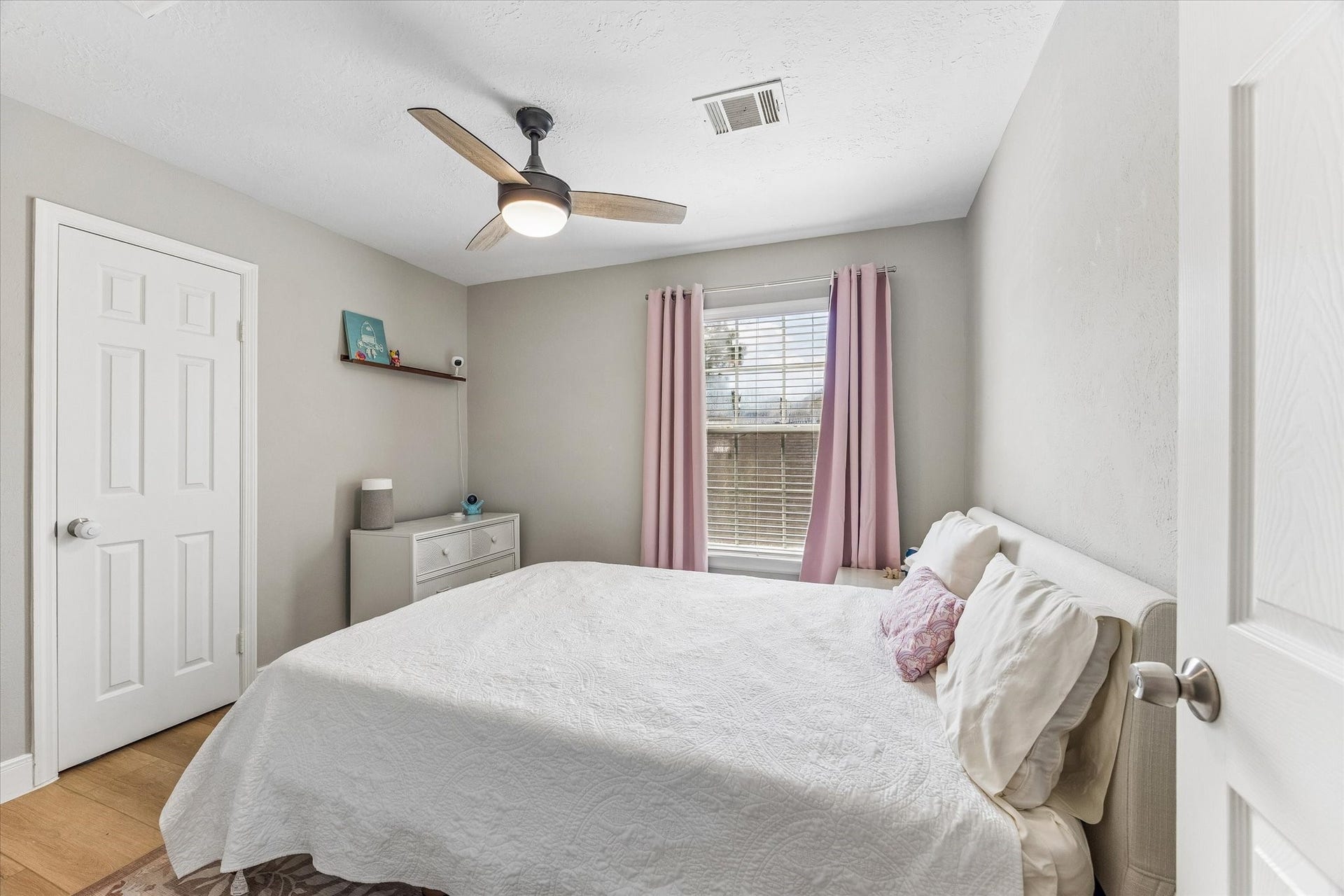 1043 Danforth Drive, Houston, TX 77062 Cozy bedroom with soft, neutral tones, featuring a comfortable bed, modern ceiling fan, and ample natural light from a window adorned with pink curtains.