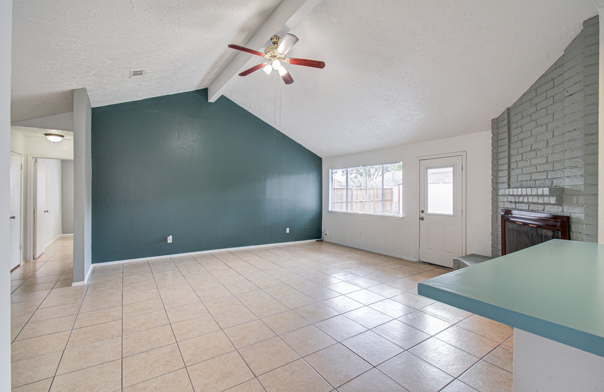 7319 Skybright Lane, Houston, TX 77095 Spacious living area with high ceilings, a ceiling fan, and abundant natural light from large windows. The room features tile flooring, a cozy brick fireplace, and a vibrant accent wall.