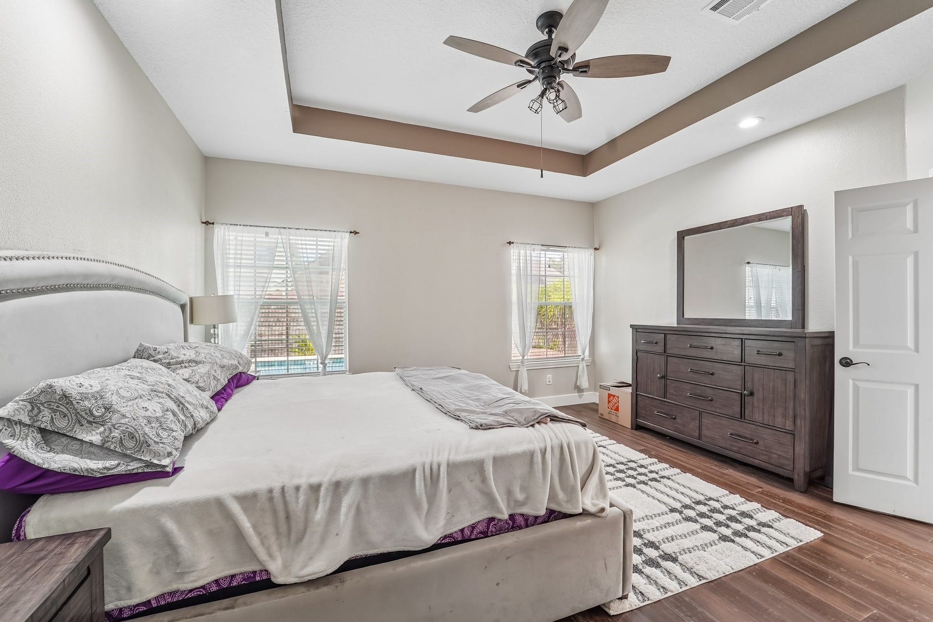 Spacious bedroom with a stylish tray ceiling, elegant ceiling fan, and abundant natural light from two large windows.