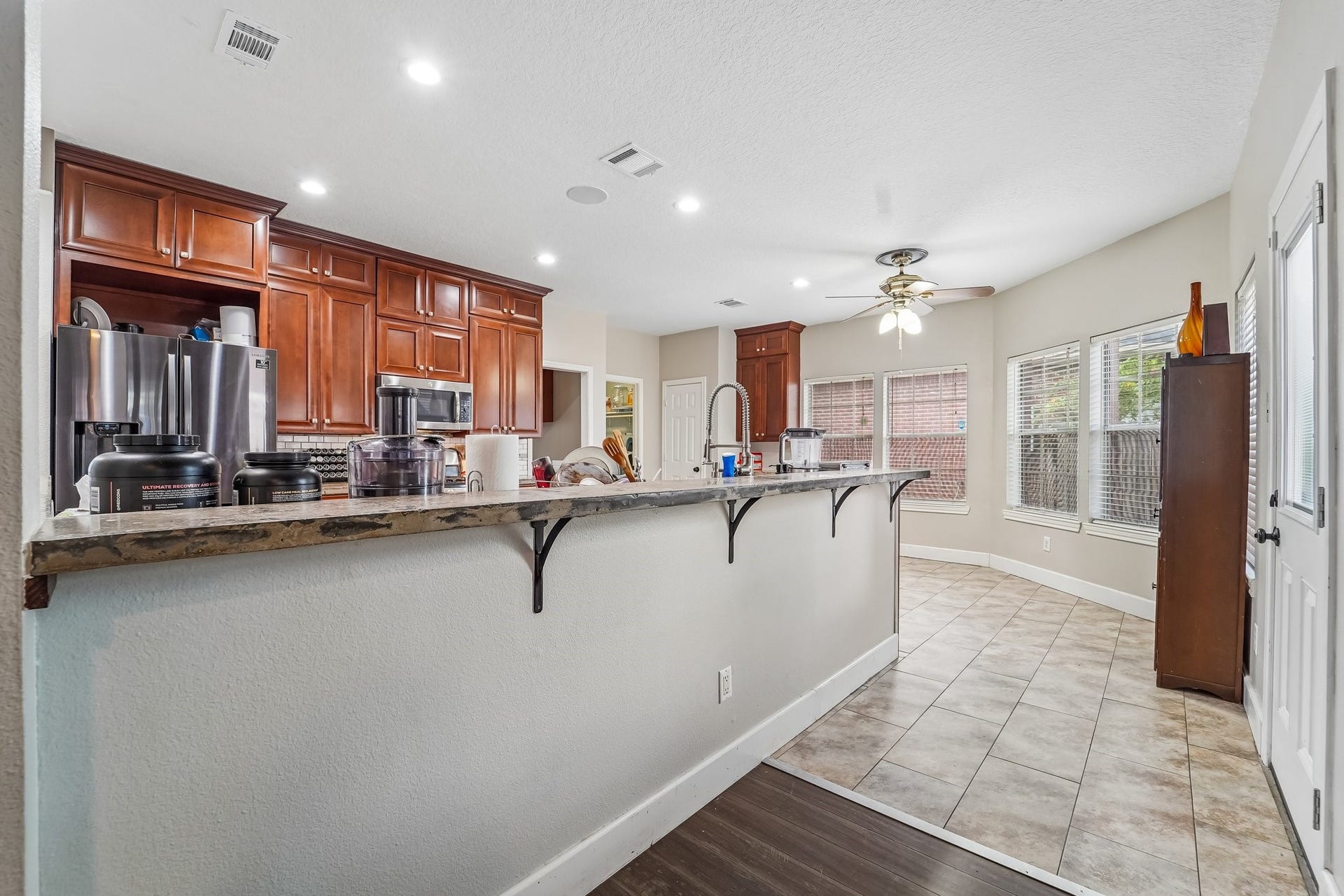 Spacious kitchen with rich wooden cabinetry, modern stainless steel appliances, and a large breakfast bar overlooking a bright dining area, enhanced by natural light from multiple windows.