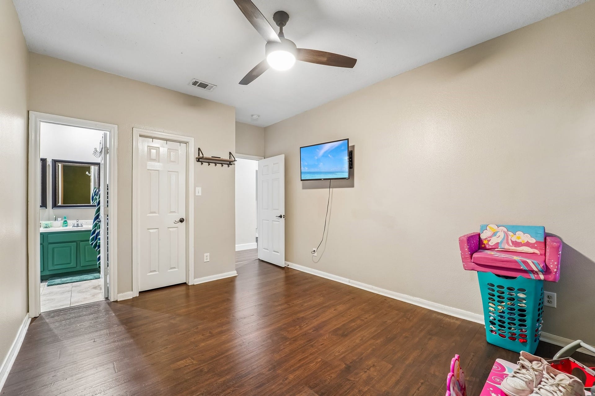 Spacious room with warm wood flooring, ceiling fan, and mounted TV, leading to a bathroom with a vibrant green vanity.