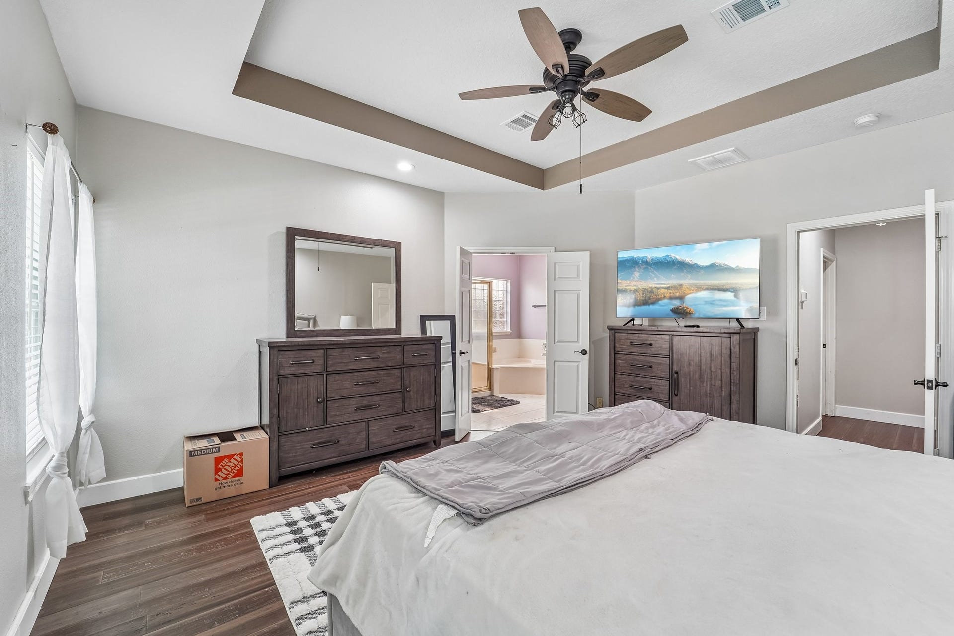 Spacious bedroom with elegant tray ceiling, featuring a ceiling fan, large dressers, and direct access to a bright en-suite bathroom.