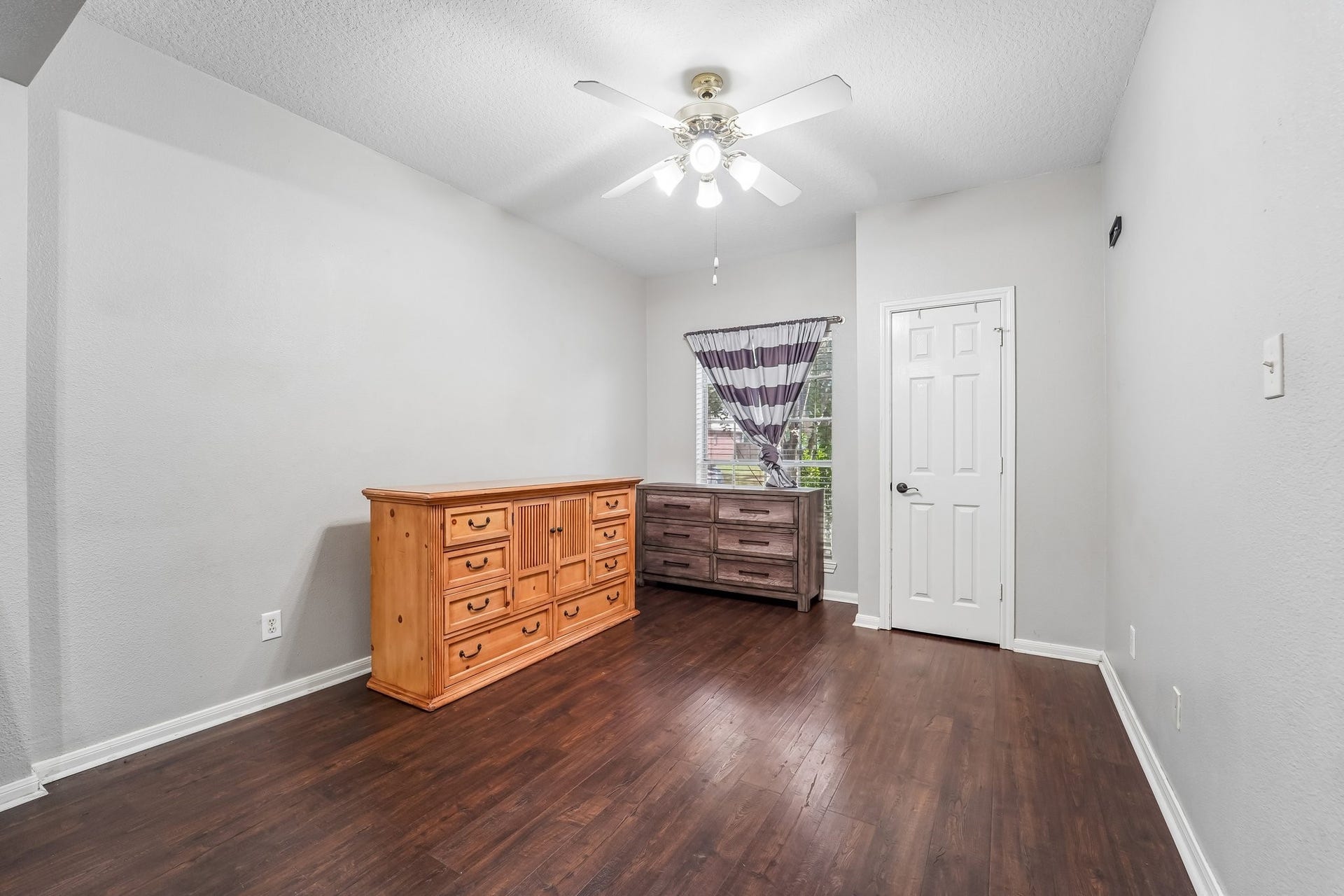 Bright room with sleek hardwood flooring, featuring a ceiling fan and ample storage with two dressers, complemented by a window for natural light.