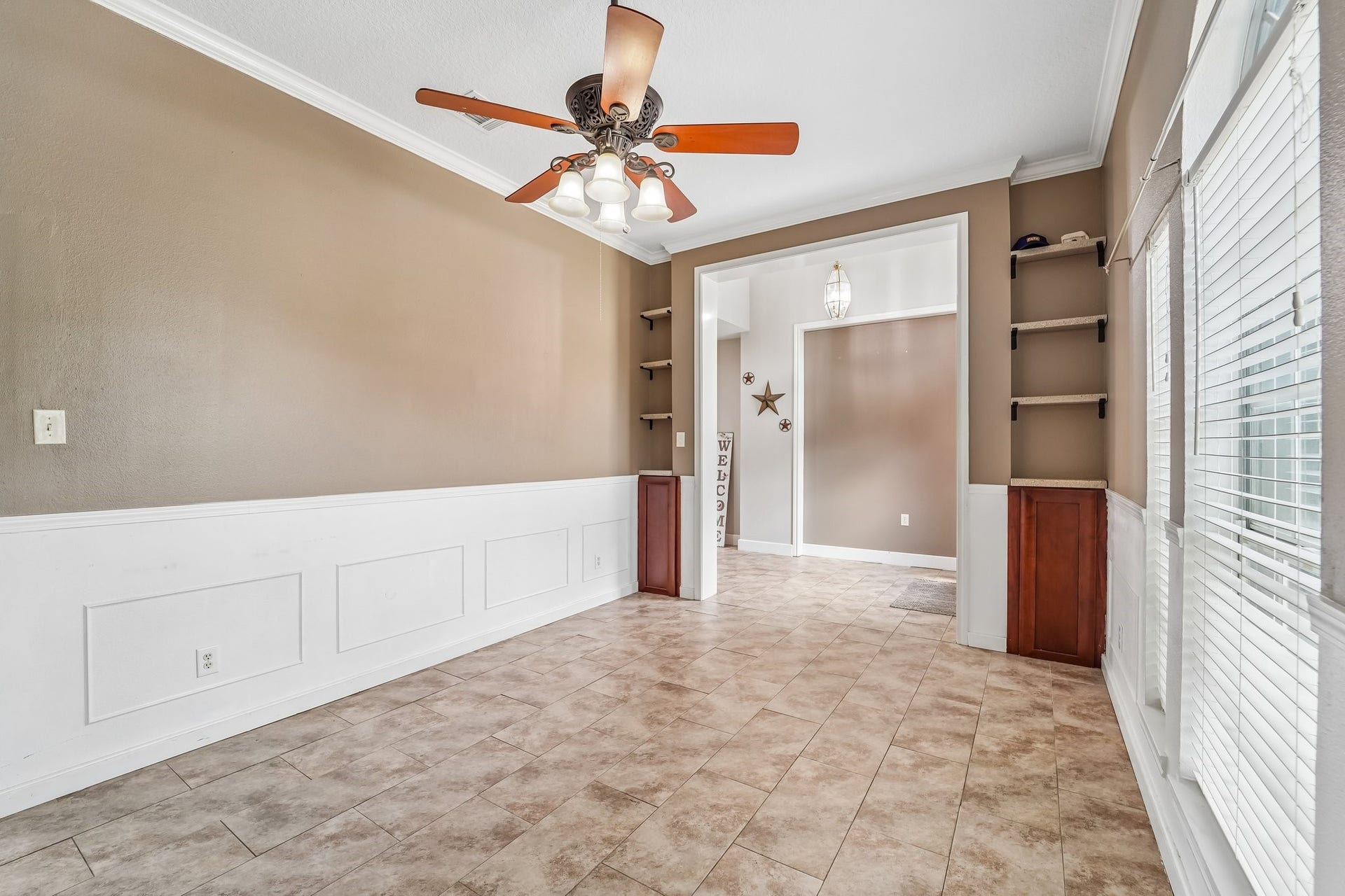Bright and inviting room featuring elegant wainscoting, a stylish ceiling fan, and built-in shelving. Ample natural light streams in through large windows, enhancing the warm, neutral tones.