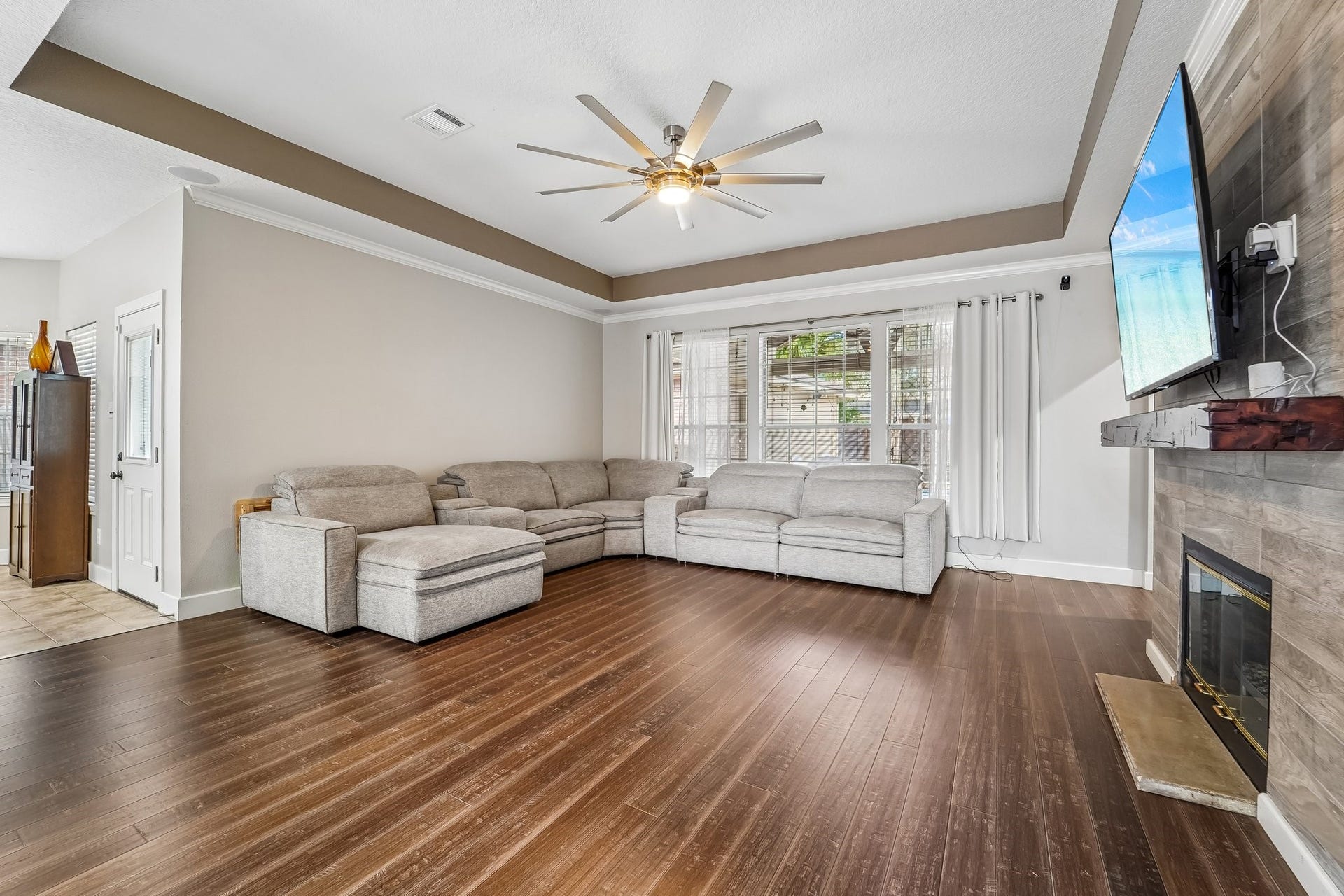 Spacious living room with elegant hardwood floors, a cozy fireplace, and a large sectional sofa, all under a stylish tray ceiling with a modern fan.