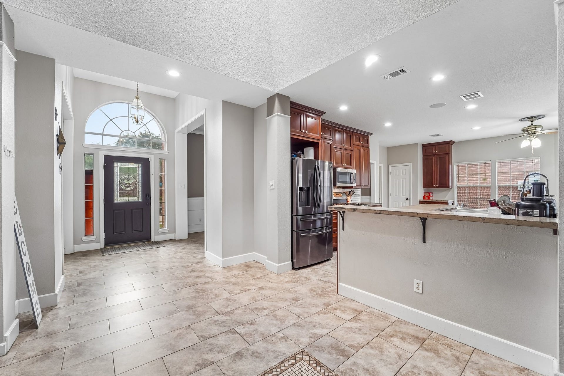 Bright and inviting entryway leads to an open kitchen featuring rich wood cabinetry, modern stainless steel appliances, and a convenient breakfast bar, all enhanced by recessed lighting and a spacious layout.