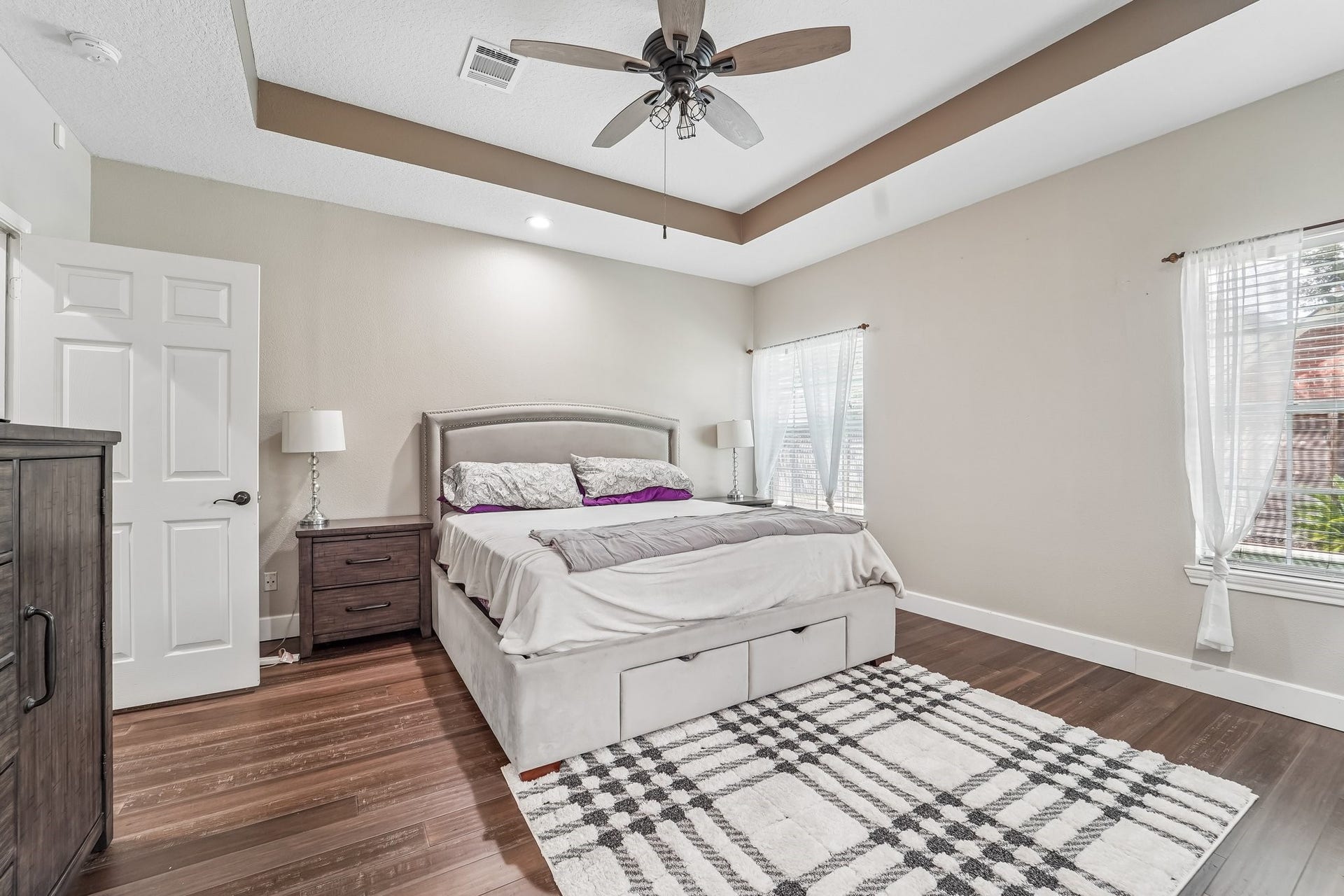 Spacious bedroom with elegant tray ceiling, abundant natural light, and warm hardwood flooring.