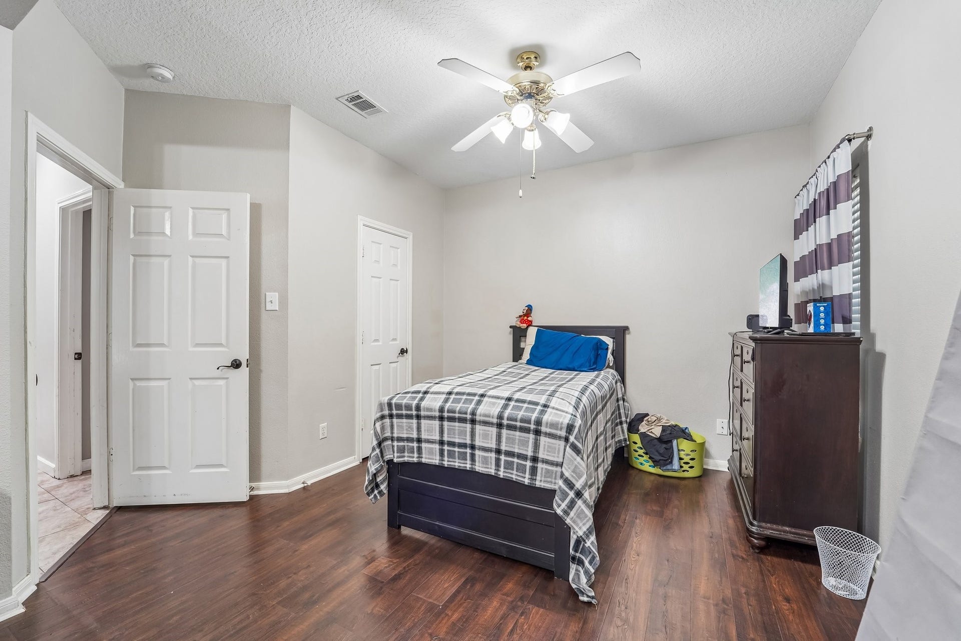 Cozy bedroom featuring warm wood flooring, a ceiling fan for comfort, and ample natural light from a window with striped curtains.
