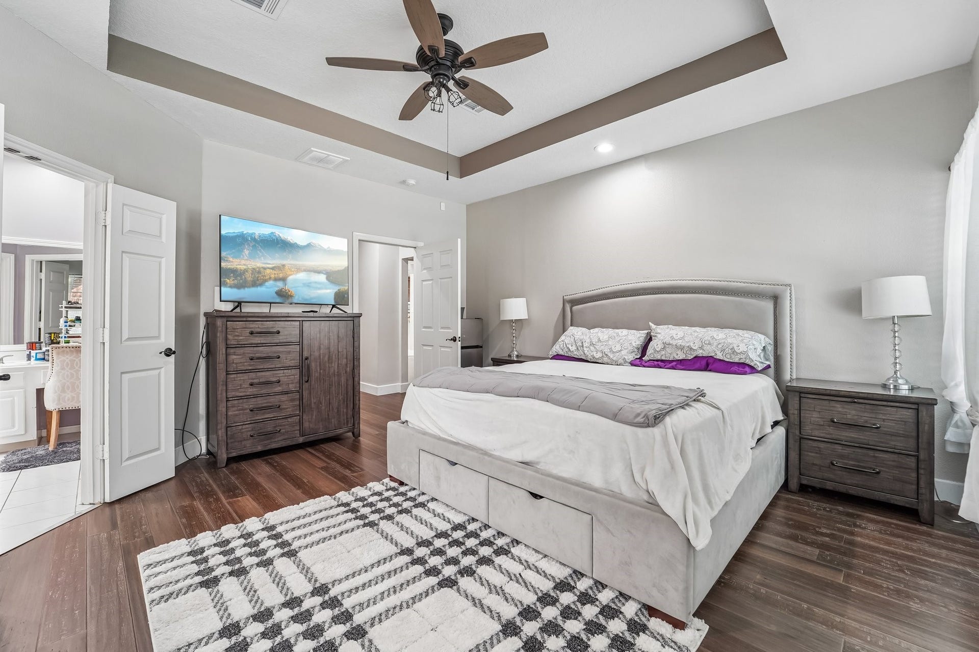 Spacious bedroom with a tray ceiling and elegant fan, featuring a plush bed, modern furniture, and abundant natural light.