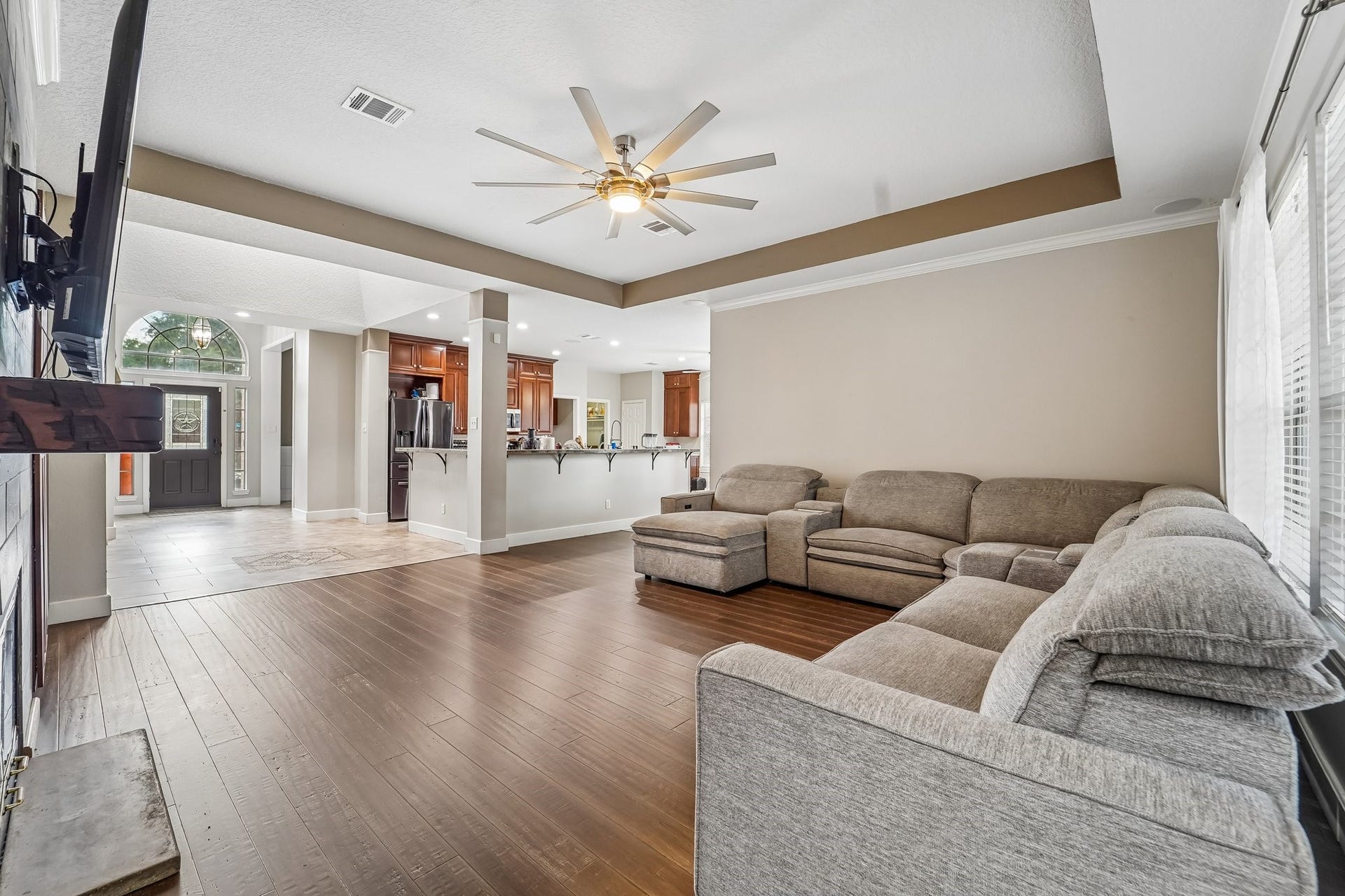 Spacious living area with an inviting open floor plan, featuring a cozy sectional sofa, rich wood flooring, and a ceiling fan, seamlessly connecting to the modern kitchen and entryway.
