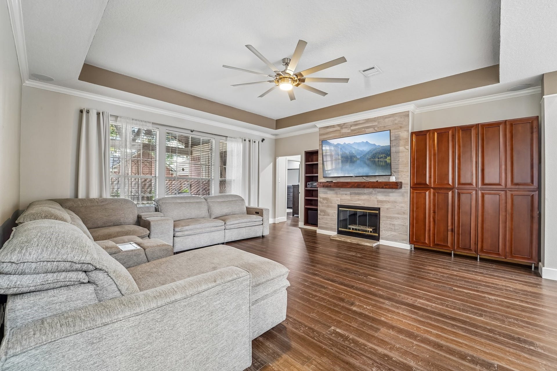 Spacious living room featuring a cozy fireplace, large sectional sofa, stylish ceiling fan, and elegant wood flooring, complemented by abundant natural light from expansive windows.