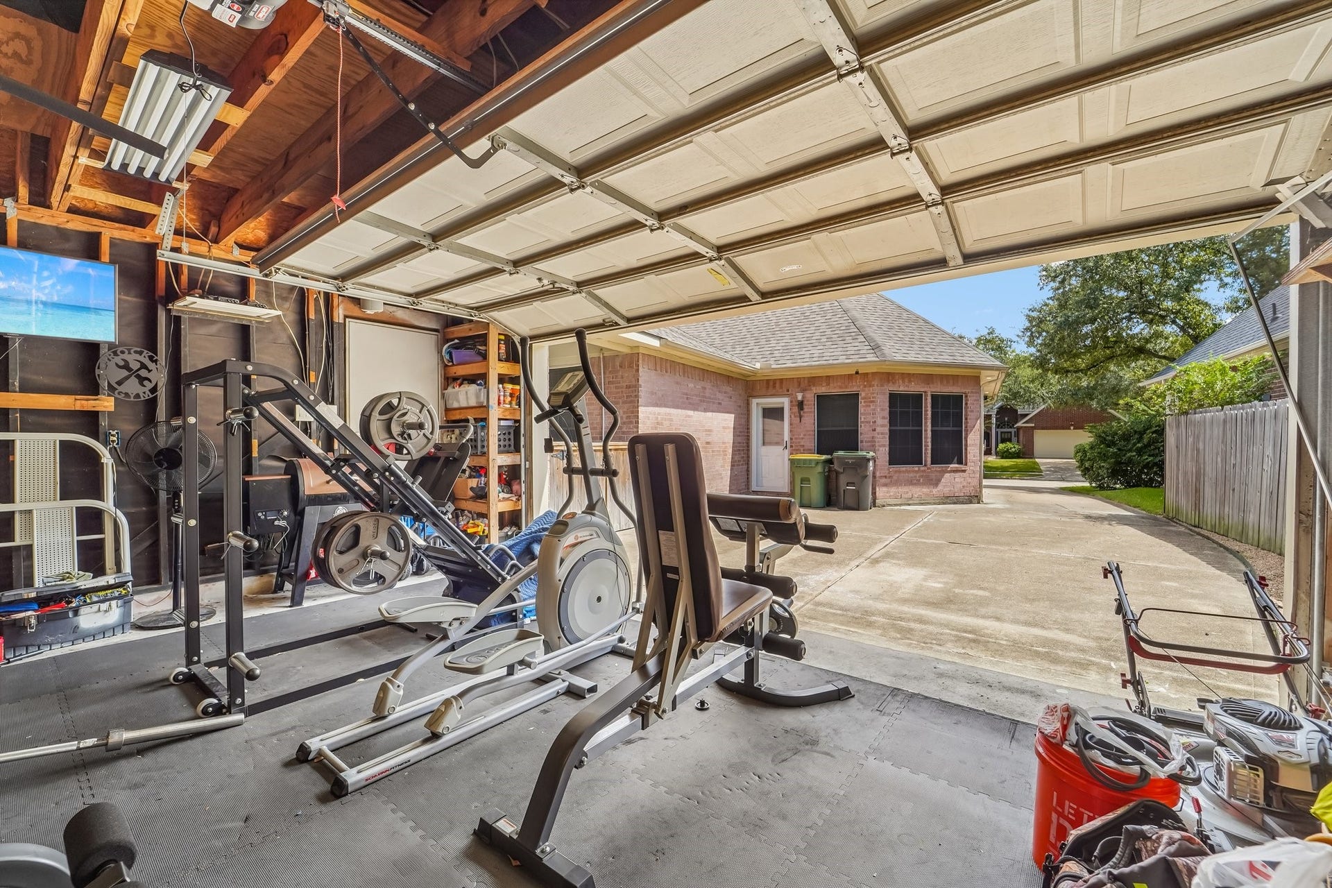 Spacious garage with home gym setup, featuring exercise equipment and ample storage. The open garage door reveals a view of the driveway leading to a charming brick house with a manicured yard.
