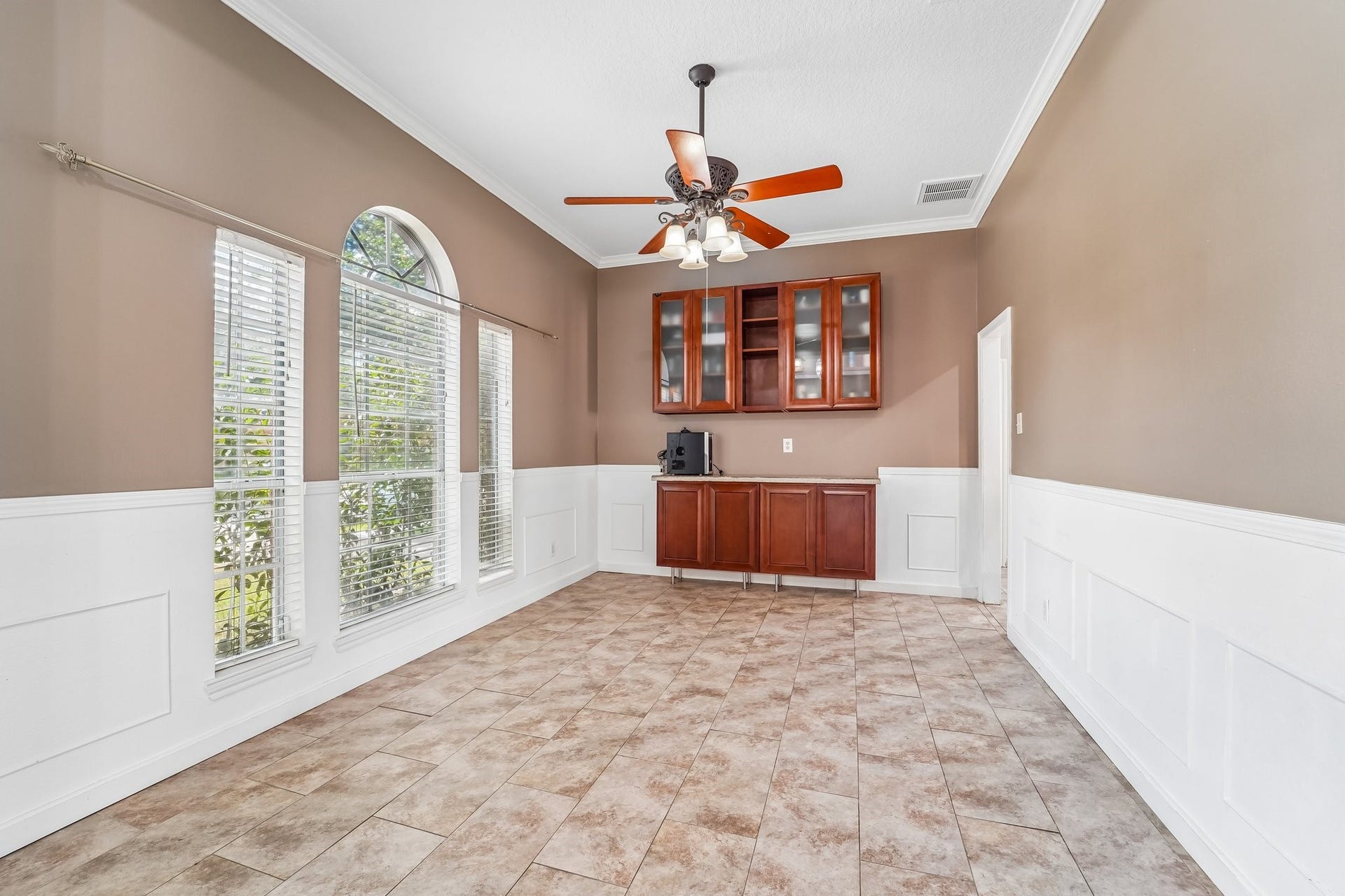 Bright and inviting room with large arched windows, elegant wainscoting, tile flooring, and a built-in wooden cabinet, perfect for a dining or multipurpose space.