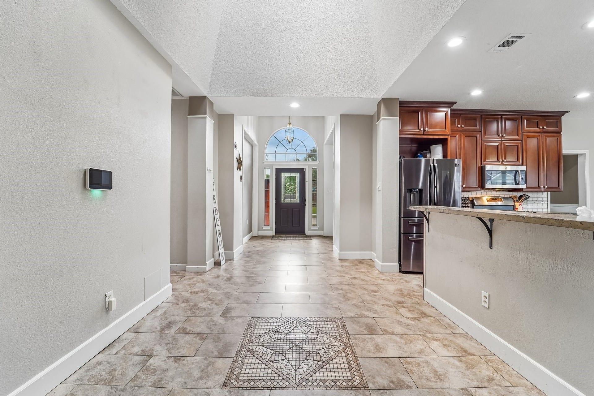 Elegant entryway with high ceilings, tile flooring, and abundant natural light, seamlessly leading into a kitchen featuring rich wooden cabinetry and modern appliances.