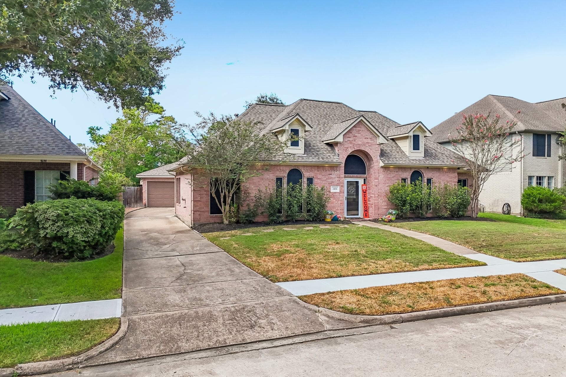 Charming brick home with a welcoming red front door, arched windows, and a manicured front lawn. A spacious driveway leads to a detached garage, surrounded by mature trees.