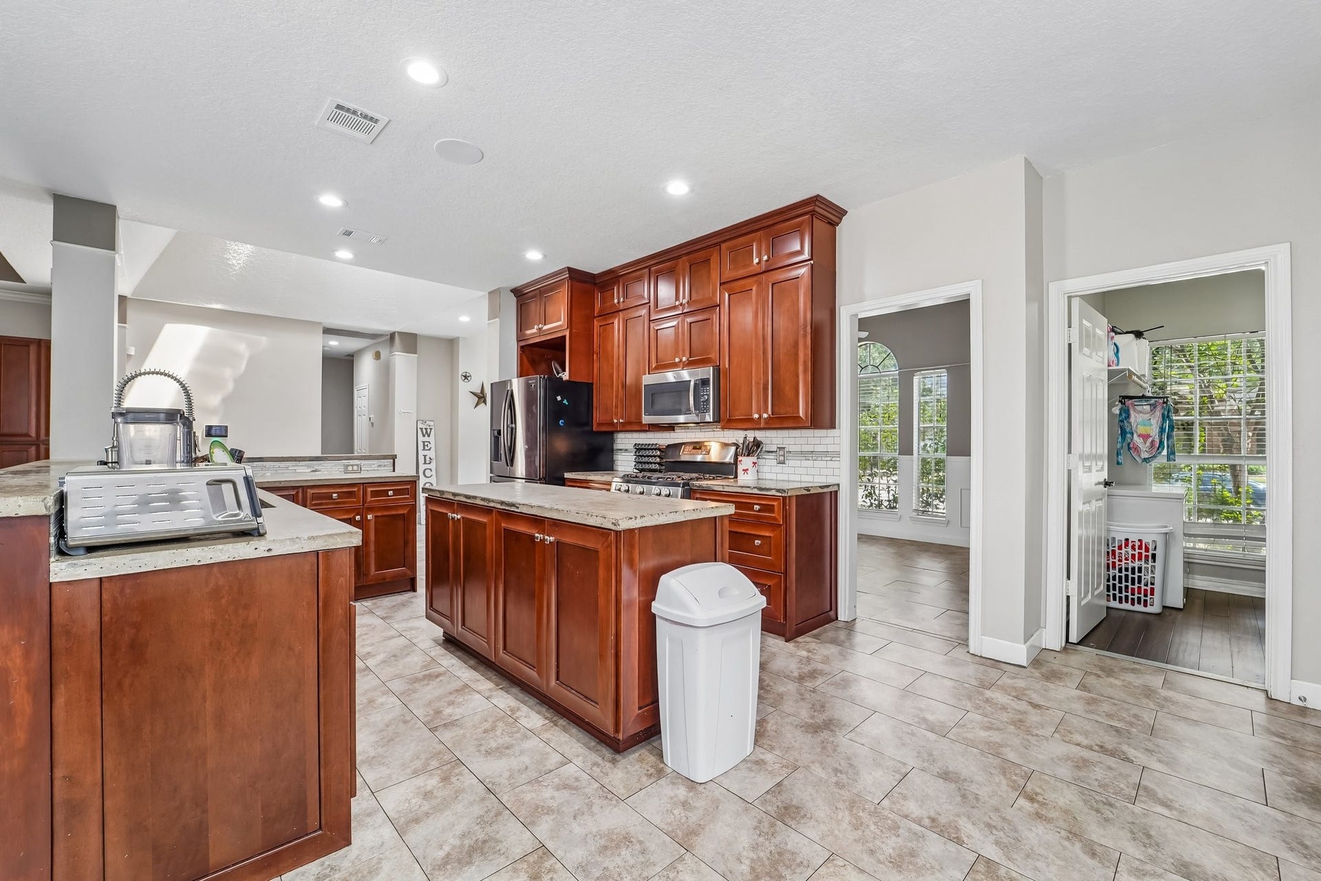 Spacious kitchen with rich wooden cabinetry, modern stainless steel appliances, and a central island, leading to a bright laundry area and sunlit room with large windows.